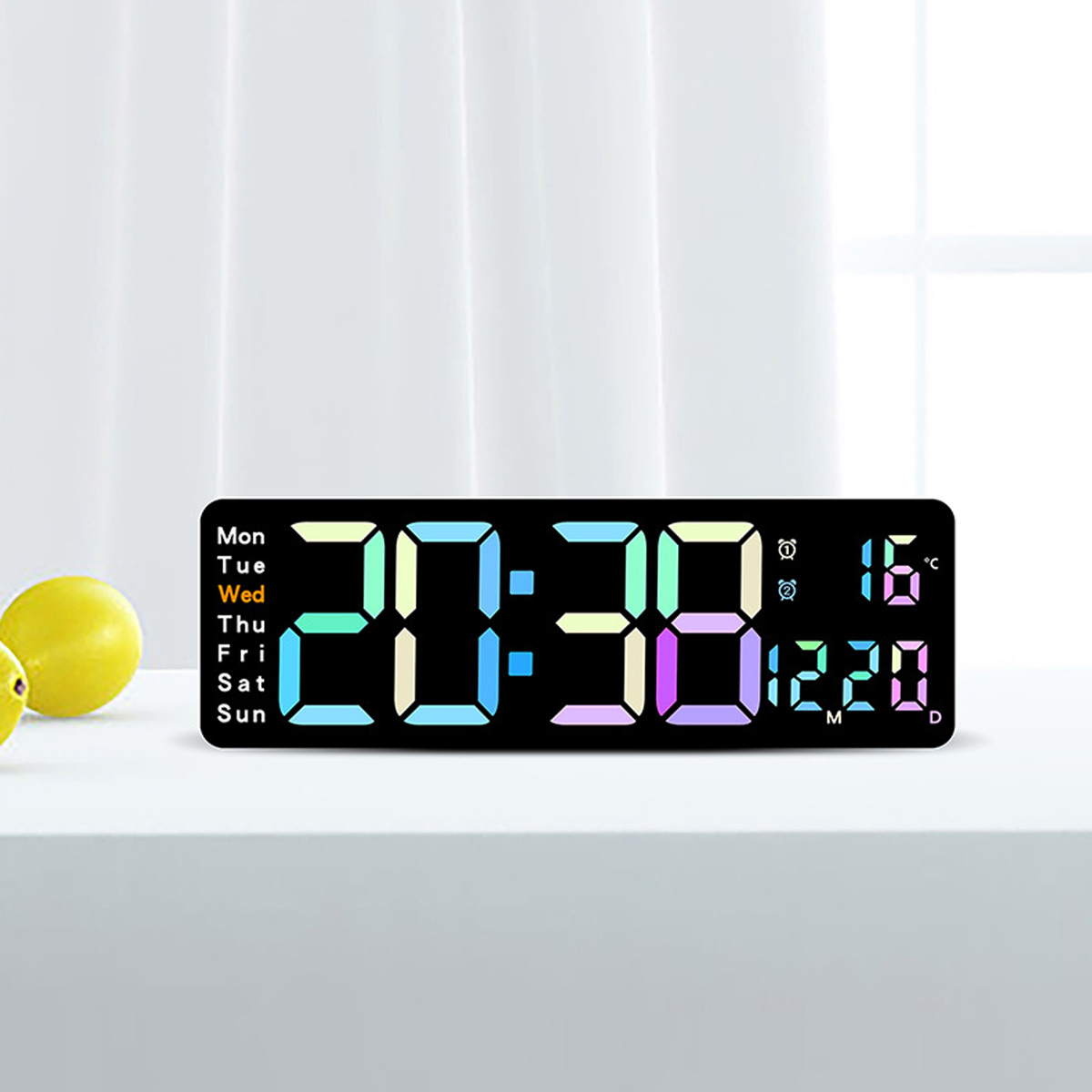 Eine Digitaluhr zeigt 20:38, 16 Grad Celsius und das Datum an. Links sind zwei gelbe Zitronen. Weißer Vorhang im Hintergrund.