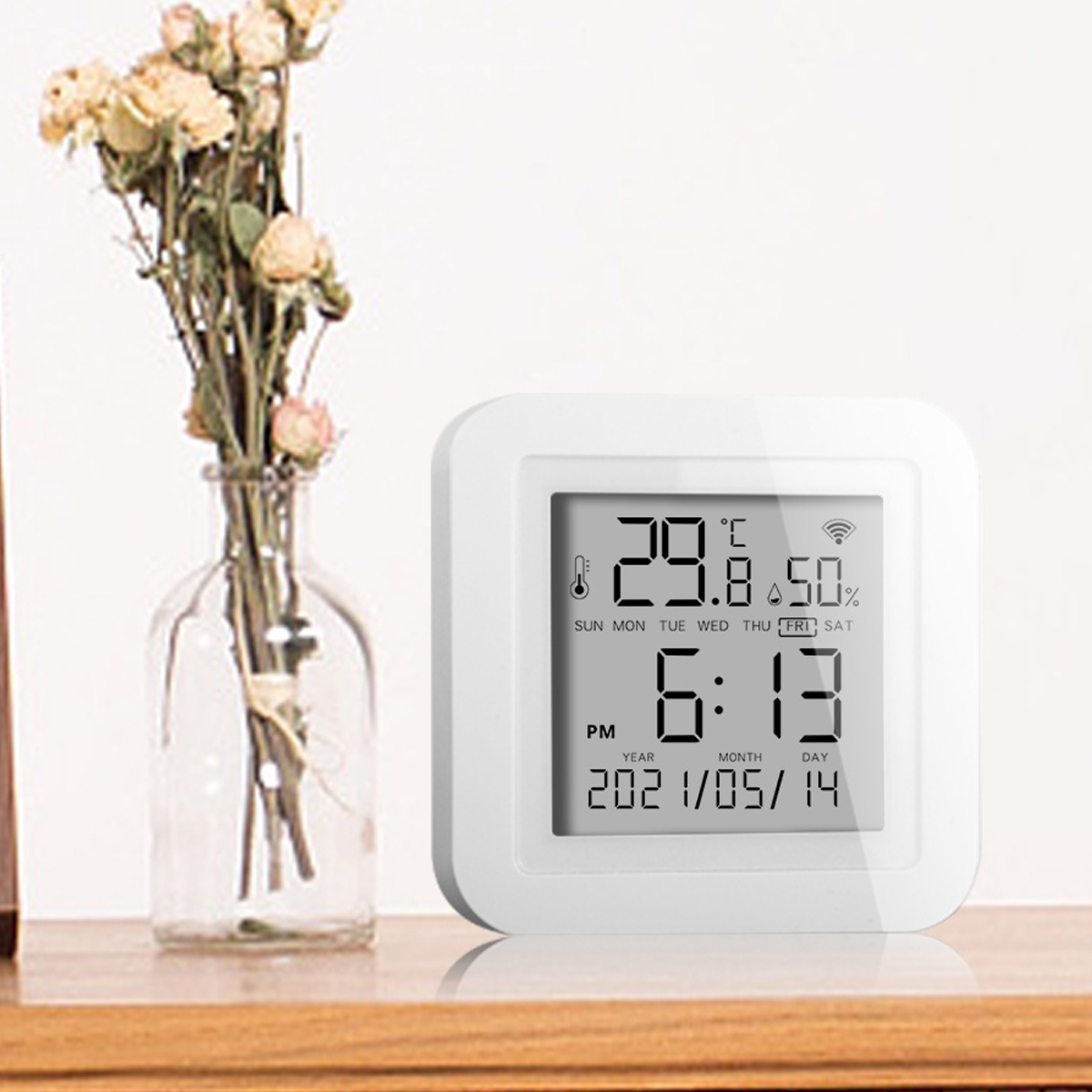Eine weiße Digitaluhr zeigt Zeit, Temperatur und Luftfeuchtigkeit an. Links steht eine Vase mit Trockenblumen auf einer Holzoberfläche.