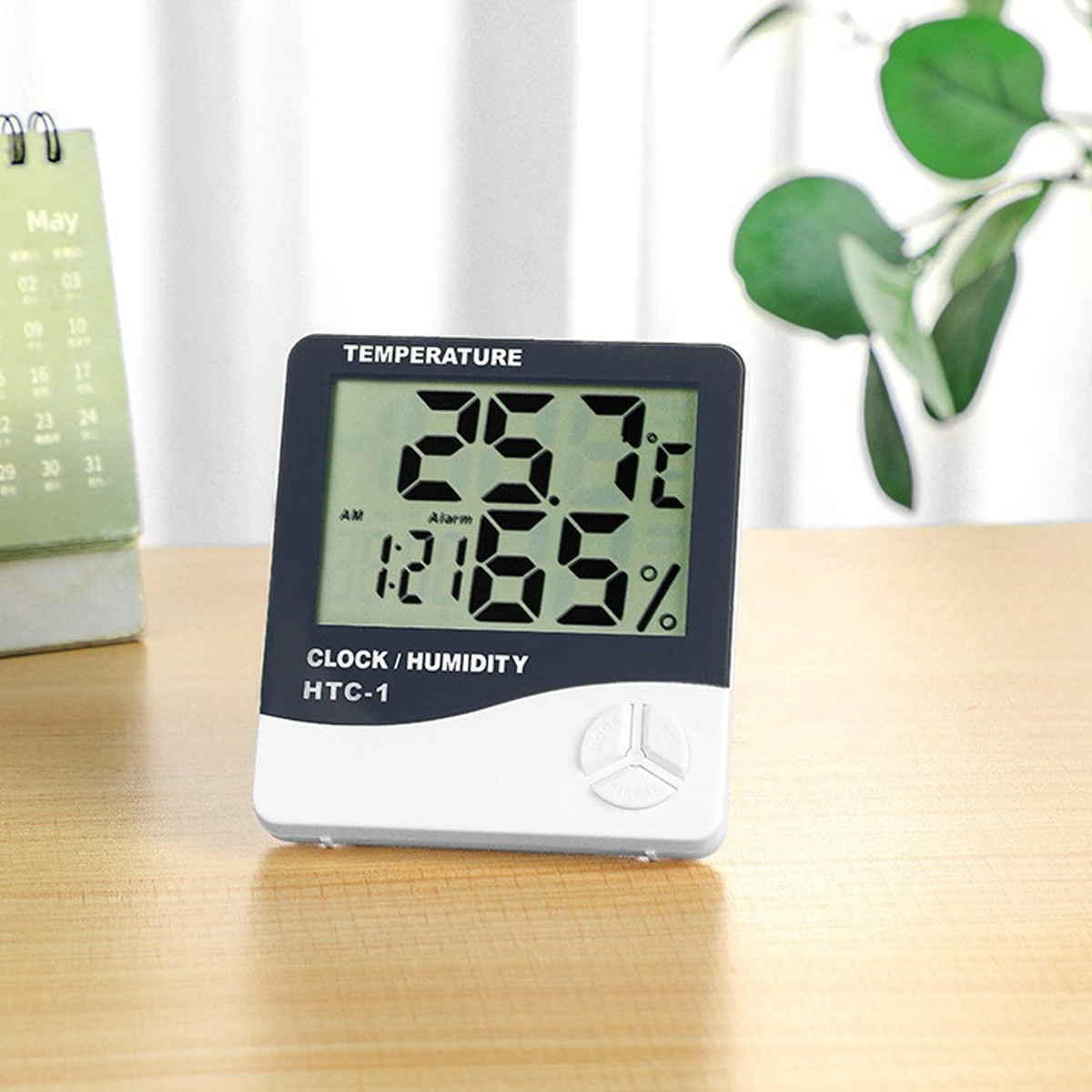 Eine digitale Uhr/Feuchtigkeitsmesser zeigt 25,7°C, 65 % und 1:21 Uhr an. Im Hintergrund befinden sich ein Kalender und eine Pflanze. Das Messgerät steht auf Holz.