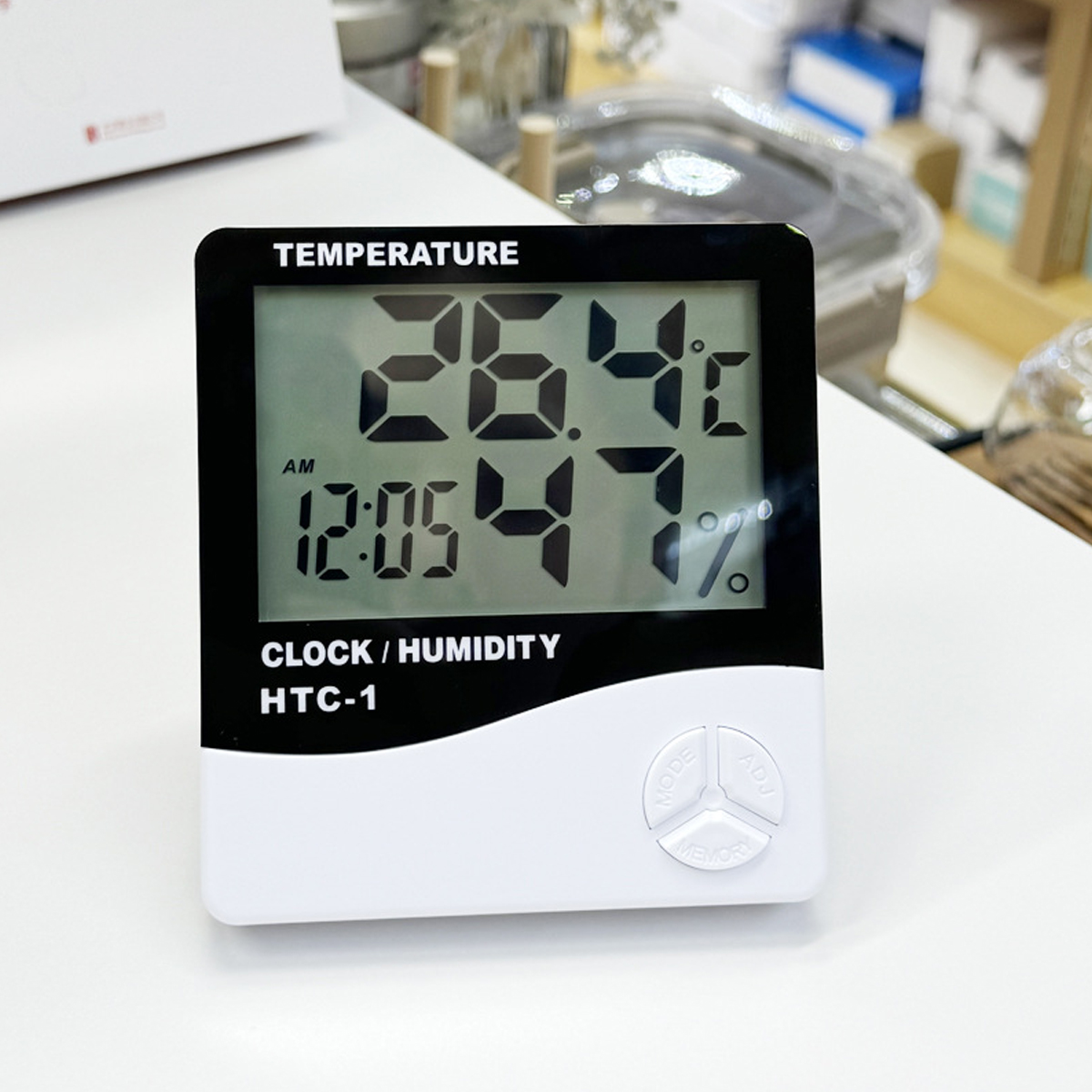 Ein digitales Uhr-/Feuchtigkeitsmessgerät zeigt 26,4°C, 47 % und 12:05 Uhr an. Das Gerät ist schwarz und weiß mit drei Tasten.