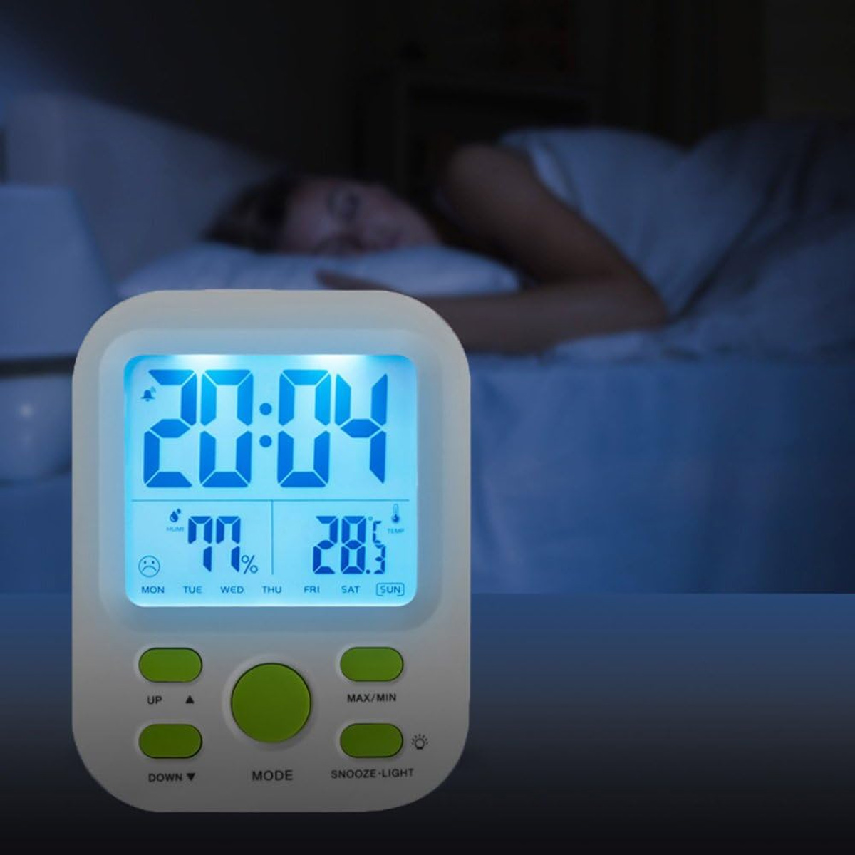 Eine Digitaluhr zeigt 20:04, 77% Luftfeuchtigkeit und 28,5°C Temperatur. Im unscharfen Hintergrund schläft eine Person. Die Uhr hat Knöpfe.