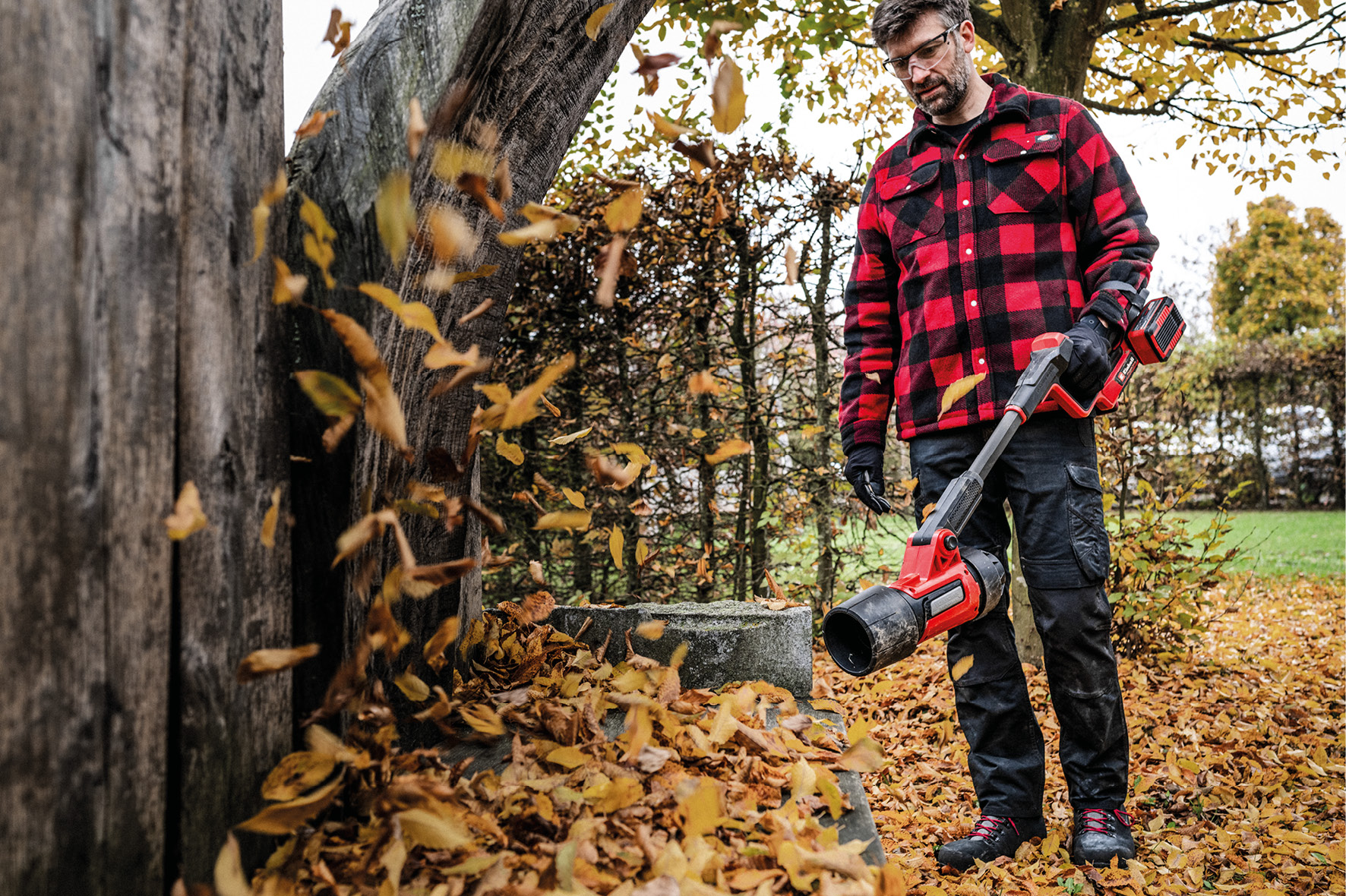 Un homme en chemise à carreaux rouge utilise un souffleur de feuilles. Les feuilles volent. Le sol est couvert de feuilles mortes.