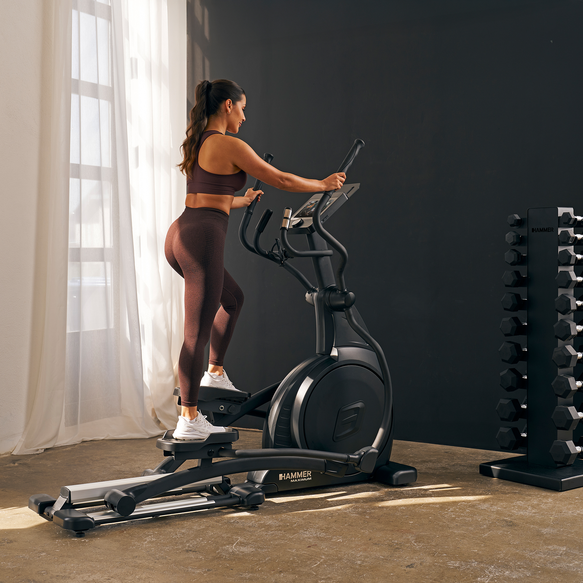 Eine Frau trainiert auf einem Crosstrainer. Sie trägt braune Trainingskleidung. Neben ihr steht ein Hantelständer.