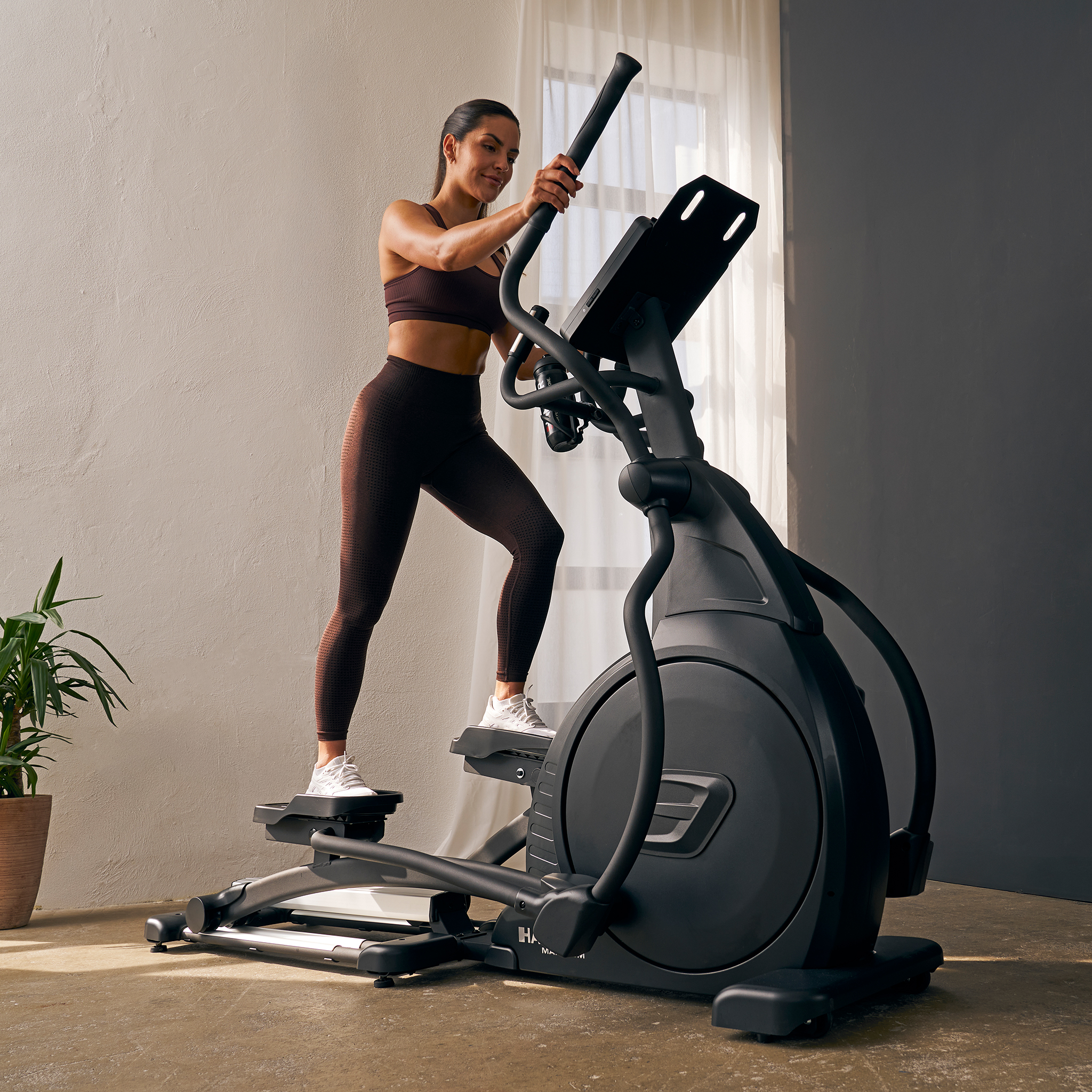 Eine Frau trainiert drinnen auf einem Ellipsentrainer. Sie trägt Sportkleidung. Die Maschine ist schwarz und die Wände sind grau.