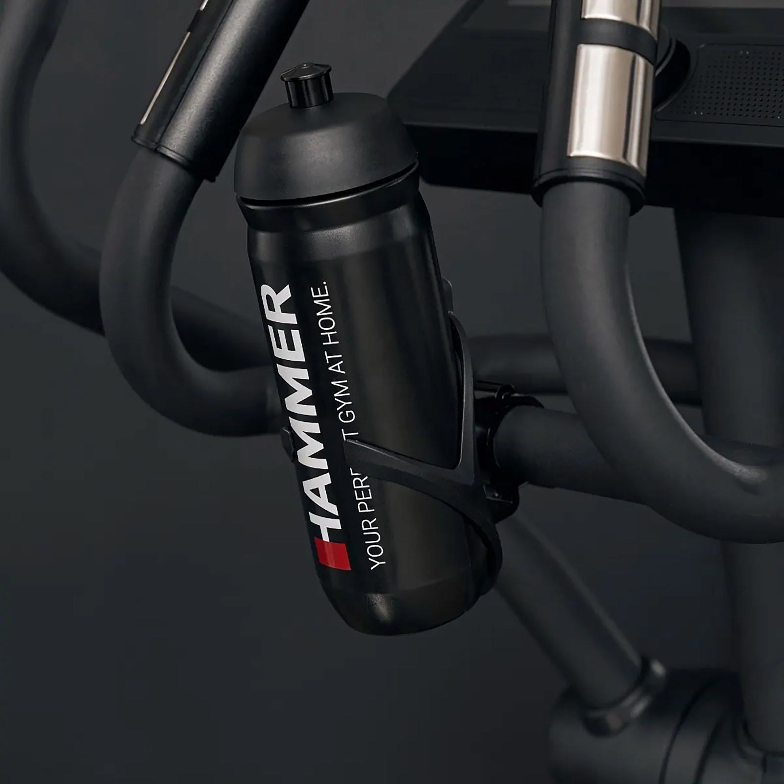 Eine schwarze Wasserflasche mit 'HAMMER'-Logo in einem Halter an einem schwarzen Trainingsgerät. Dunkelgrauer Hintergrund.