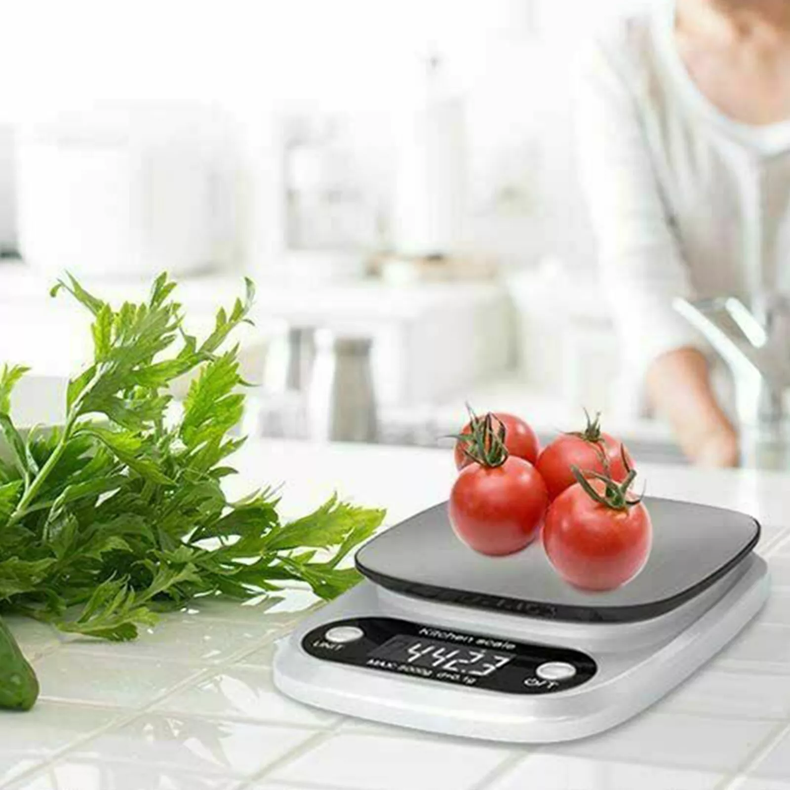 Eine Küchenwaage mit vier roten Tomaten darauf. Grüne Kräuter sind links.