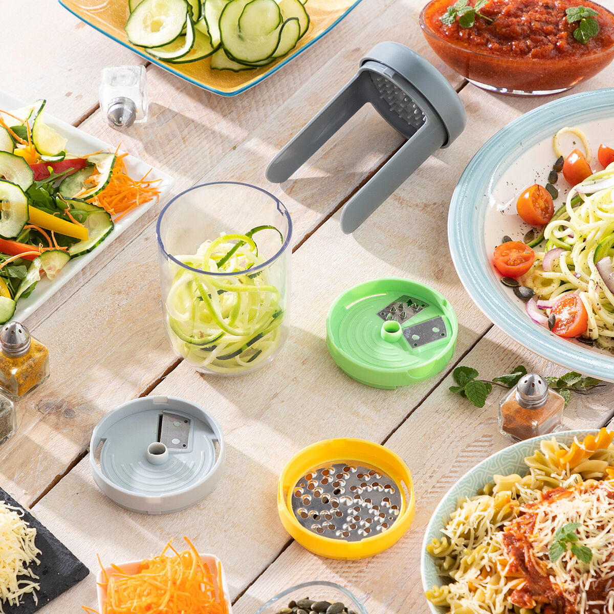 Vista dall'alto di vari alimenti, tra cui spaghetti di zucchine, pasta e verdure, con utensili da cucina su un tavolo di legno.
