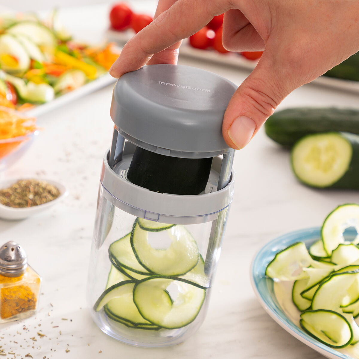 Una mano tiene un tagliaverdure grigio InnovaGoods. Fette di cetriolo sono all'interno del contenitore trasparente. Altri ingredienti circondano il tagliaverdure.
