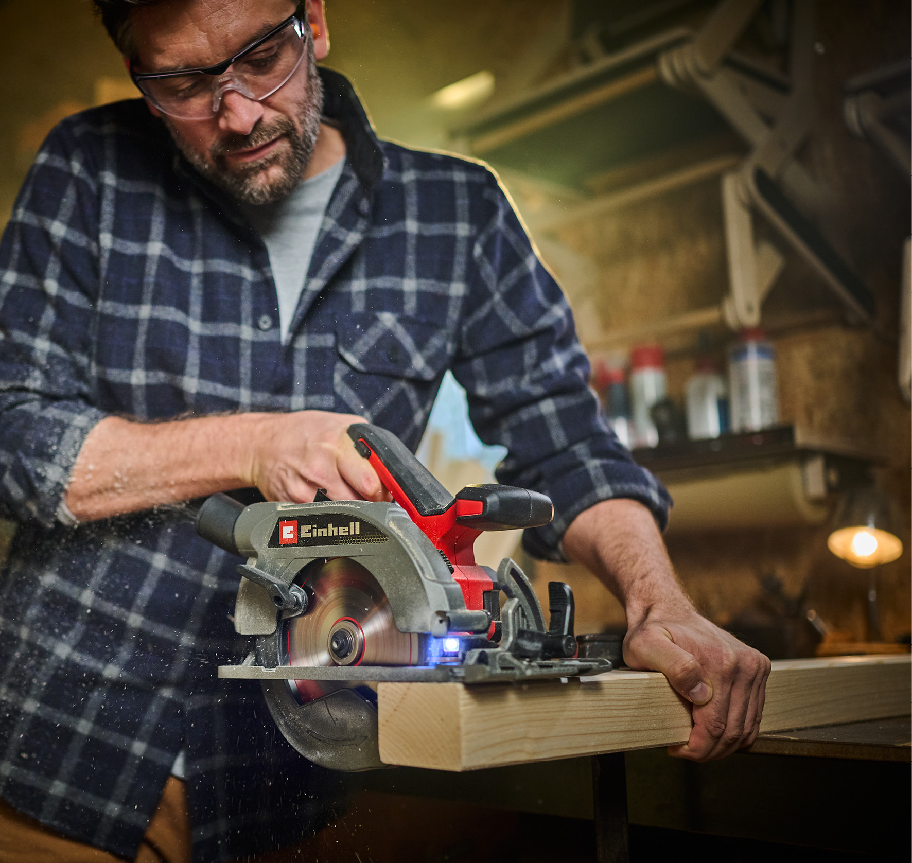 Ein Mann mit Schutzbrille schneidet mit einer Kreissäge ein Holzbrett in einer Werkstatt, Sägemehl fliegt.