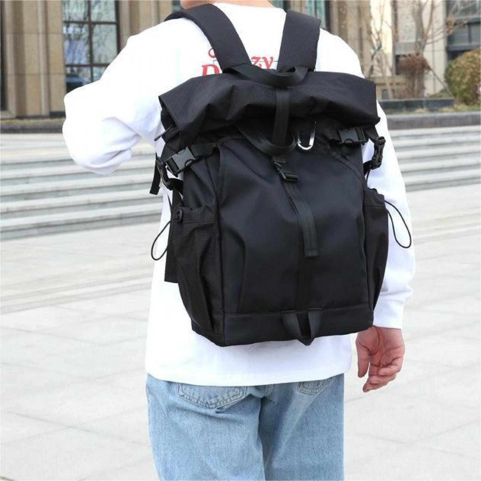 Eine Person trägt einen schwarzen Rucksack mit Riemen und Schnallen. Sie tragen ein weißes Hemd und blaue Jeans. Hintergrund ist ein Gebäude.