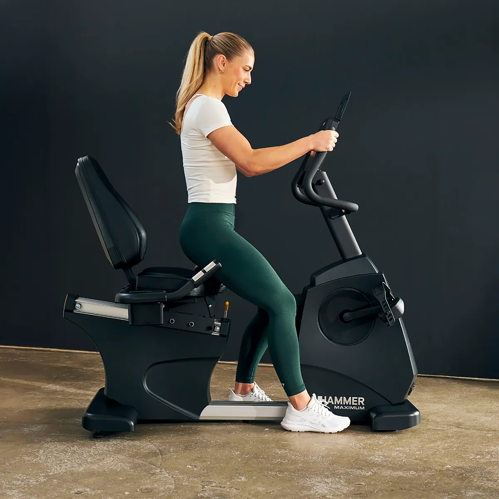 Eine Frau auf einem Heimtrainer. Sie trägt ein weißes Hemd und grüne Leggings und trainiert drinnen. Das Fahrrad ist schwarz mit der Marke HAMMER.