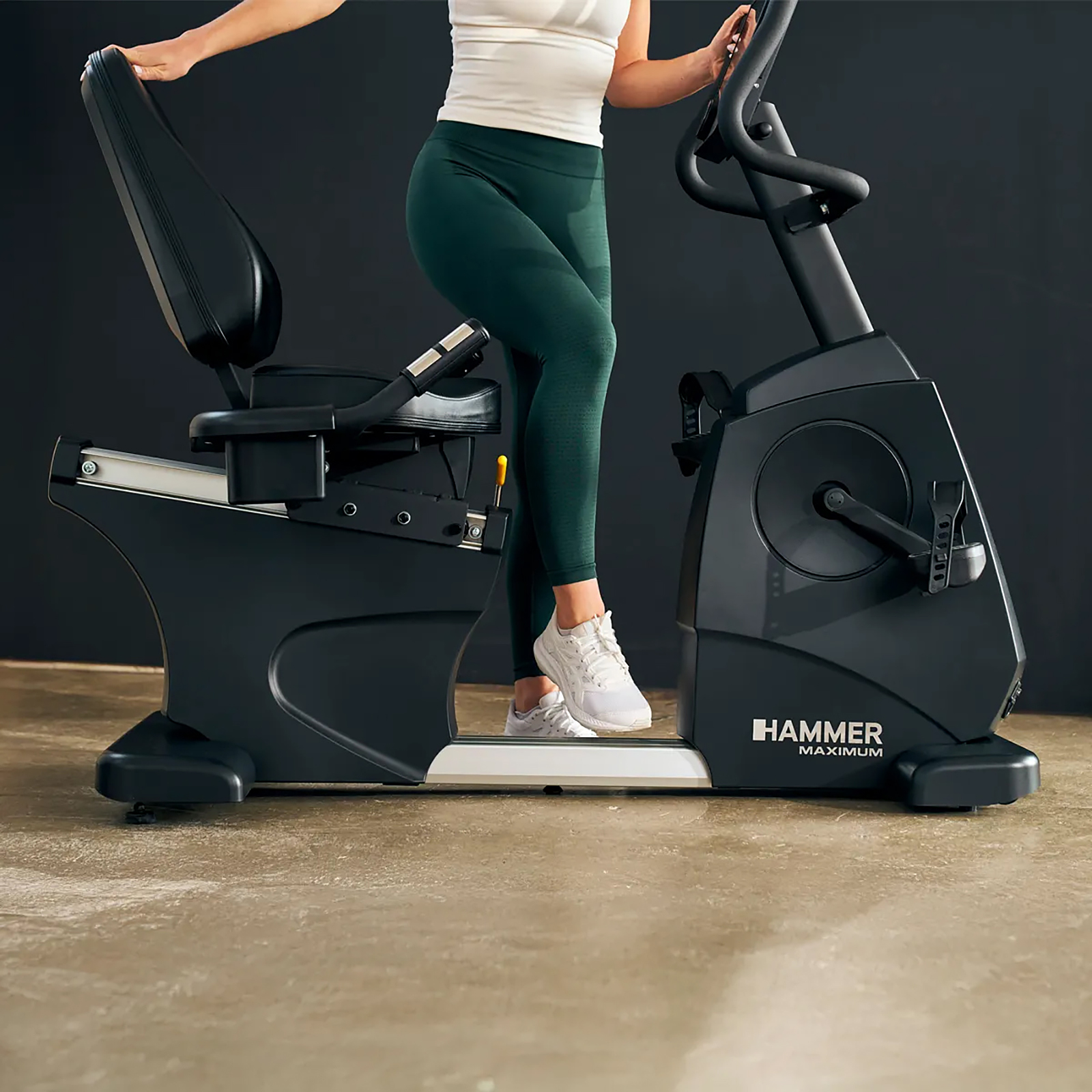 Eine Frau in Sportkleidung sitzt auf einem Heimtrainer. Das Fahrrad ist schwarz mit dem Markennamen Hammer. Der Hintergrund ist dunkel.