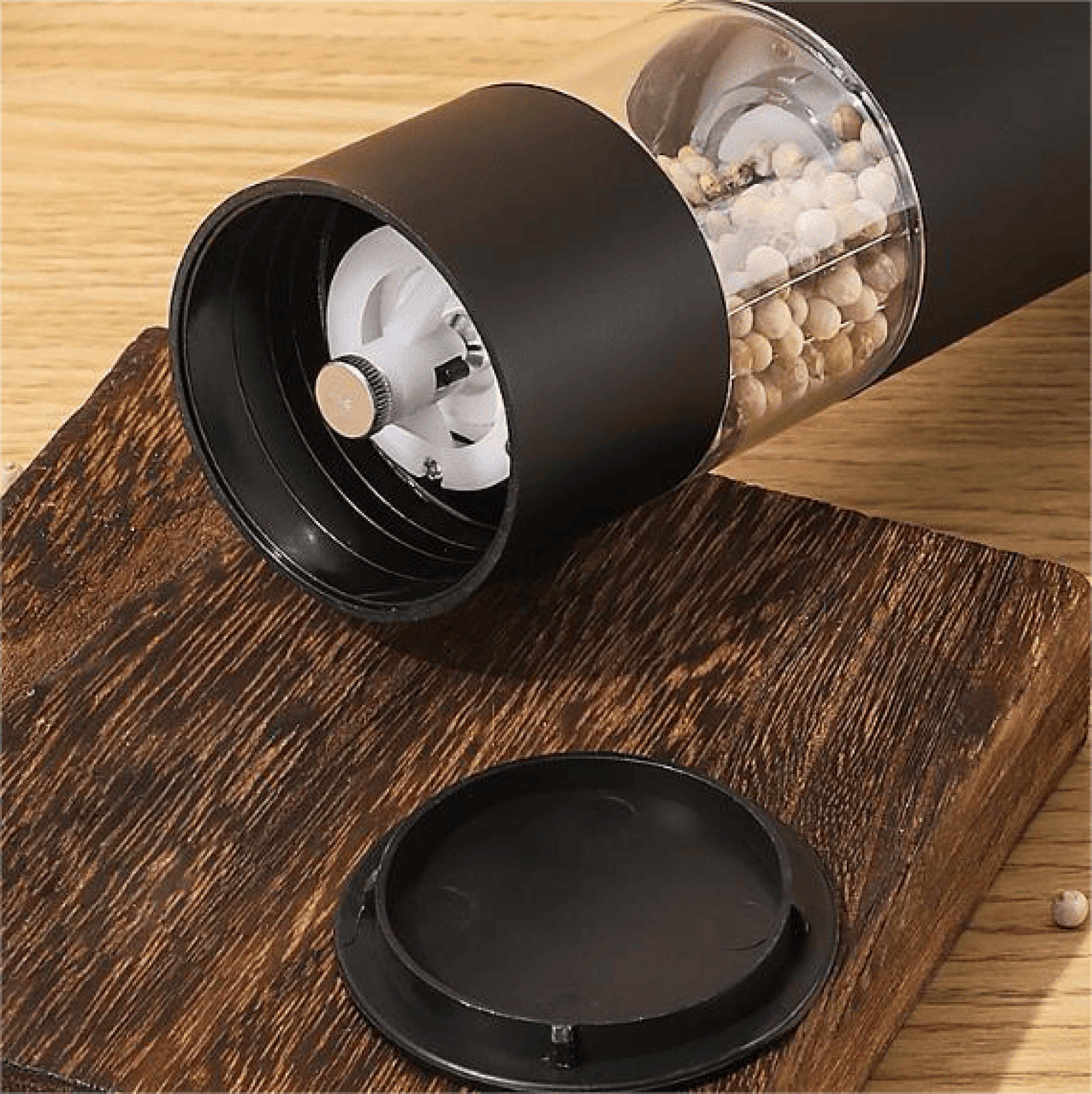 Eine schwarze und transparente Pfeffermühle mit Pfefferkörnern im Inneren, die auf einer Holzoberfläche liegt. Der Deckel ist ebenfalls sichtbar.