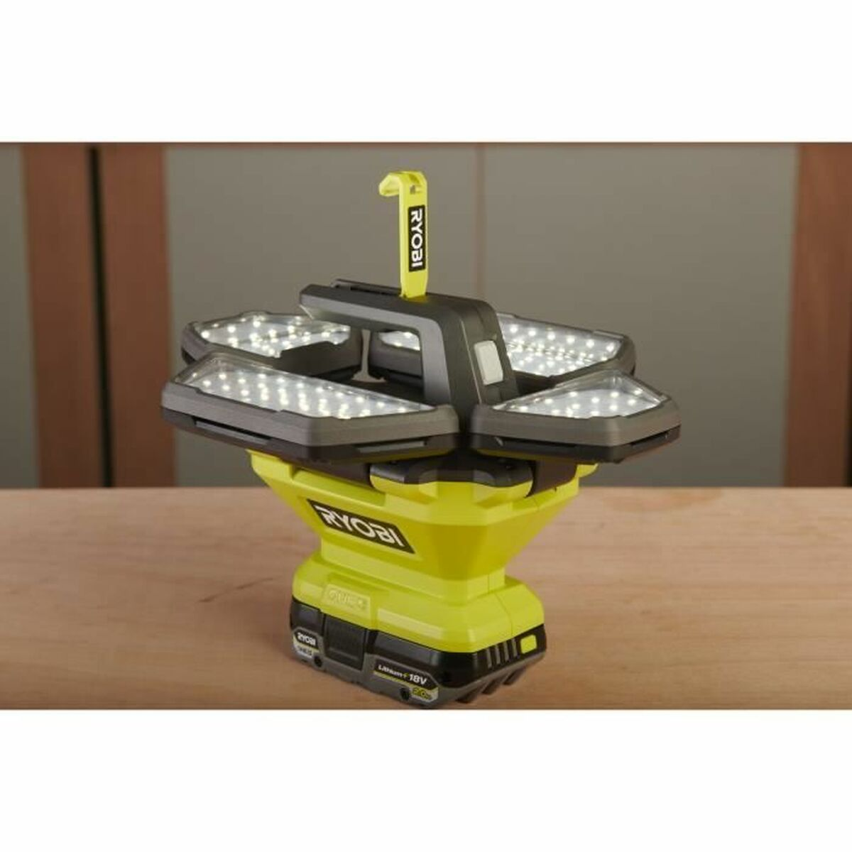 Une lampe de travail LED Ryobi. Elle possède quatre panneaux lumineux, une poignée jaune et une base noire et jaune. Elle est posée sur une surface en bois.