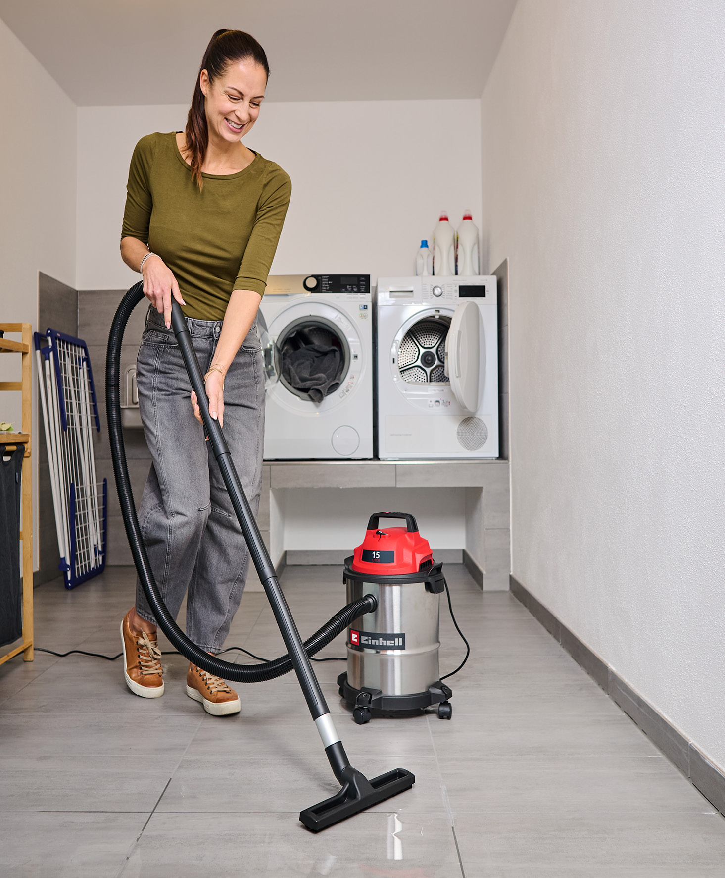 Eine Frau saugt einen gefliesten Boden mit einem rot-silbernen Staubsauger. Waschmaschinen sind im Hintergrund.