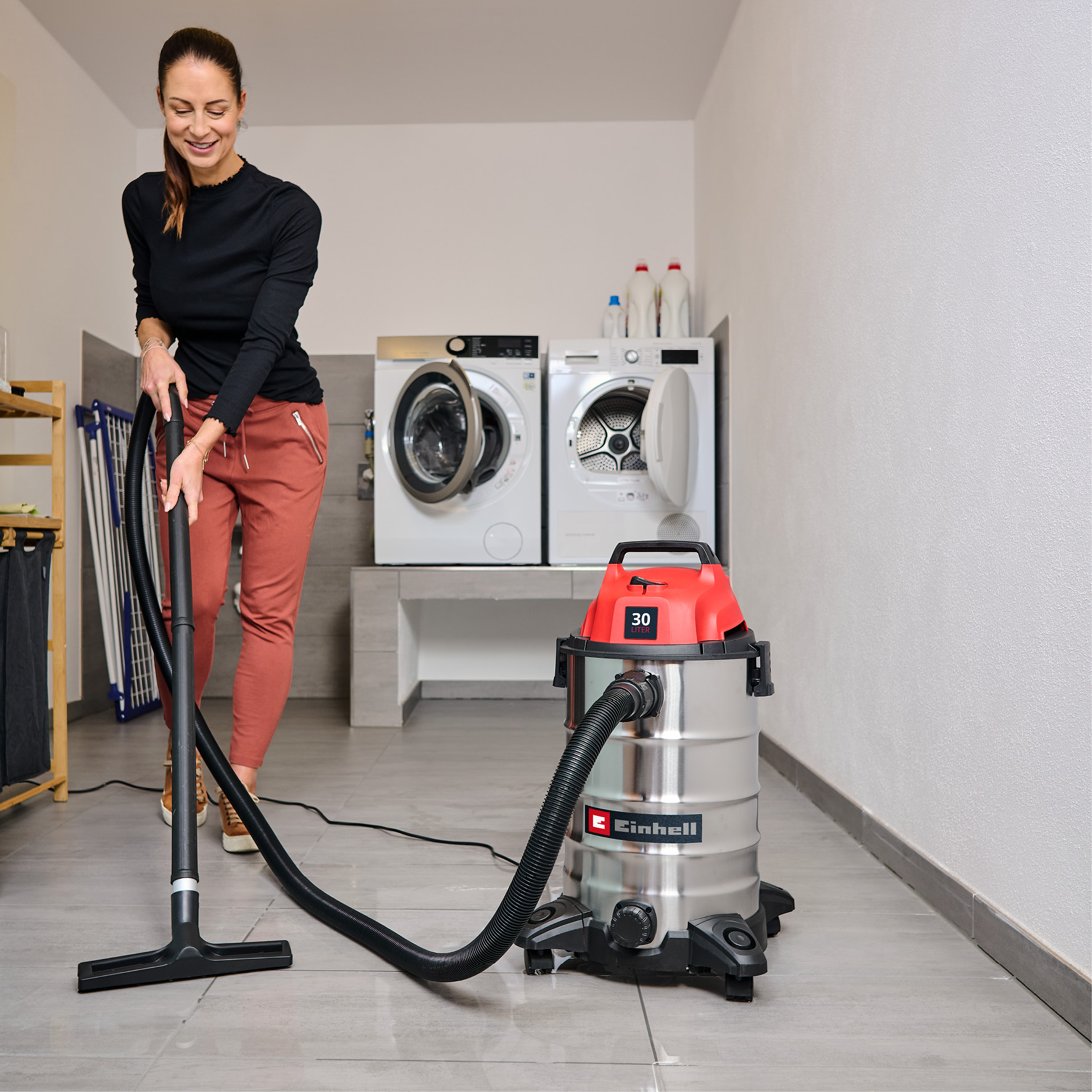 Une femme passe l'aspirateur dans une buanderie. Un aspirateur rouge et argenté est au premier plan. Des machines à laver sont en arrière-plan.