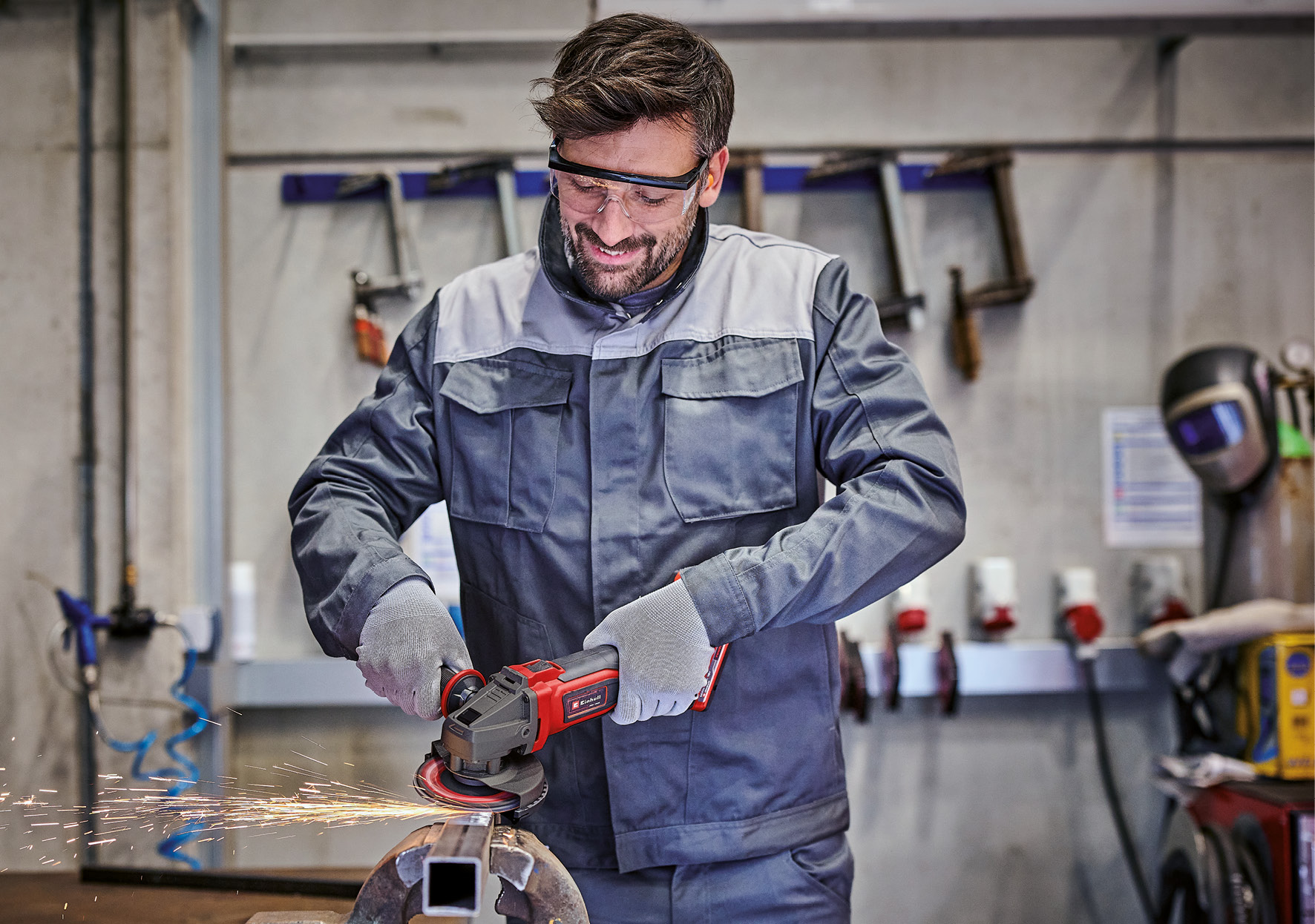 Ein Mann schleift Metall mit einem Winkelschleifer. Funken fliegen. Er trägt eine Schutzbrille, Handschuhe und einen Arbeitsanzug.