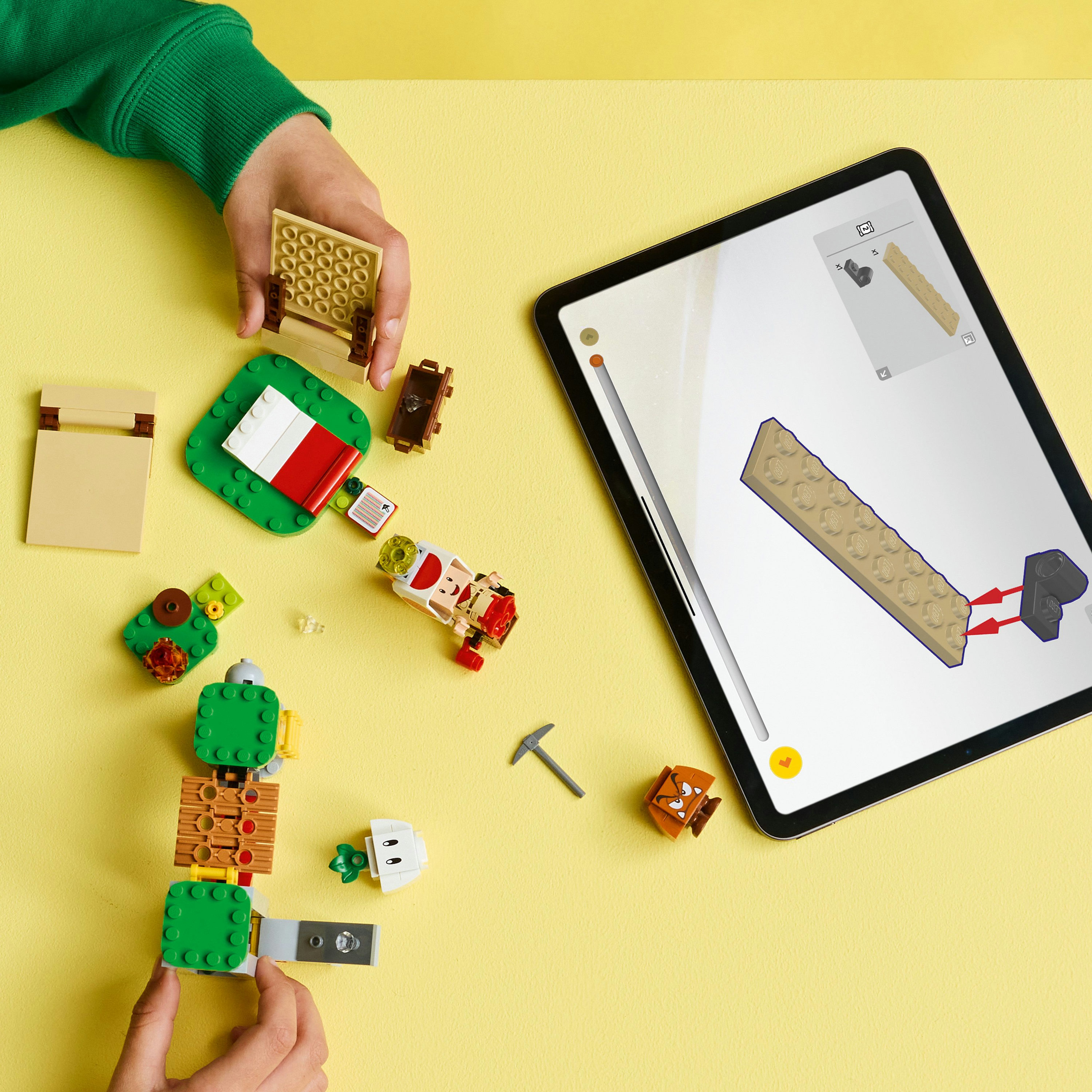 Ręce składające klocki Lego. Tablet wyświetla instrukcje. Różne klocki i figurki są rozrzucone na żółtej powierzchni.