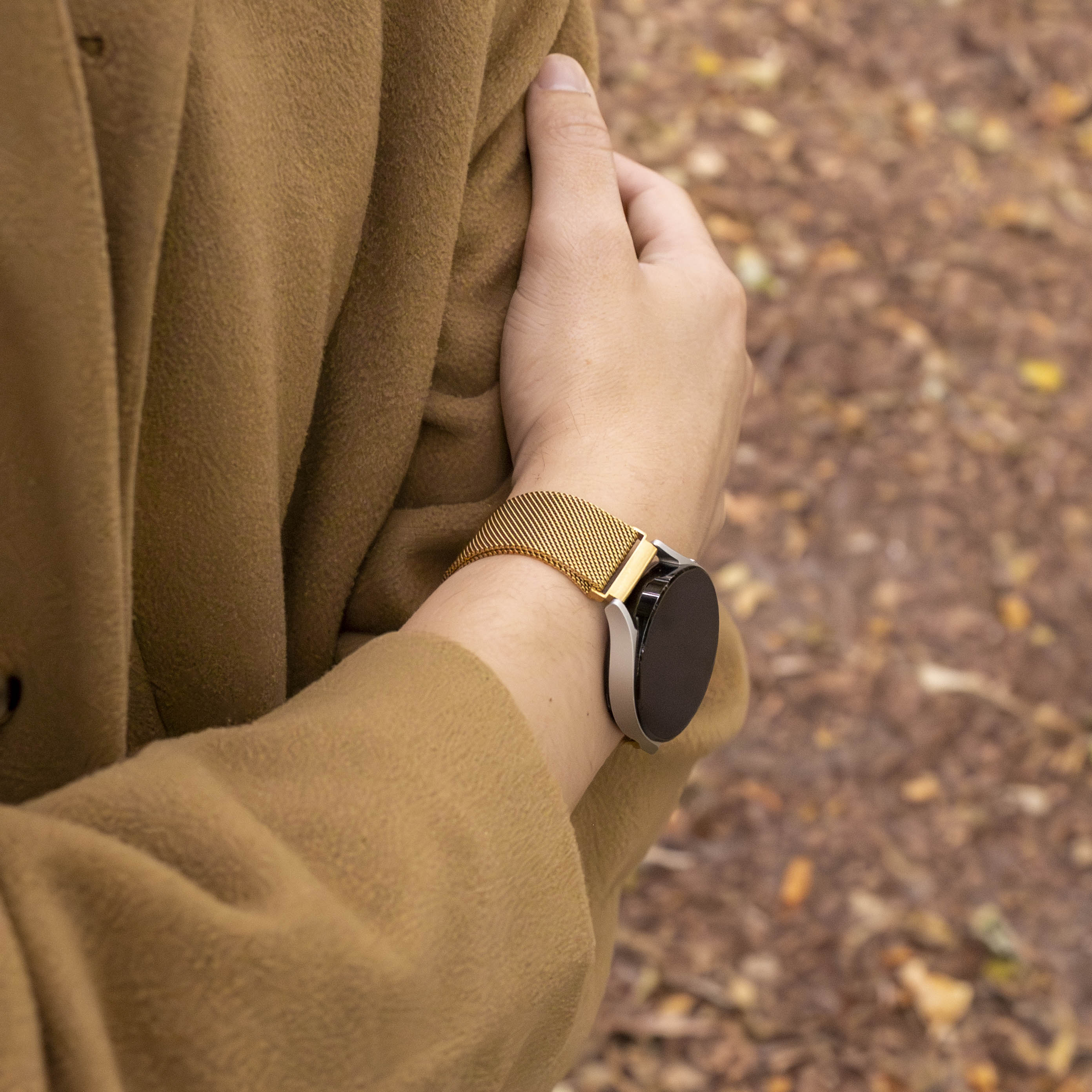 Eine Person, die eine goldene Uhr mit schwarzem Bildschirm, braunem Mantel trägt, und der Hintergrund sind verschwommene Herbstblätter.