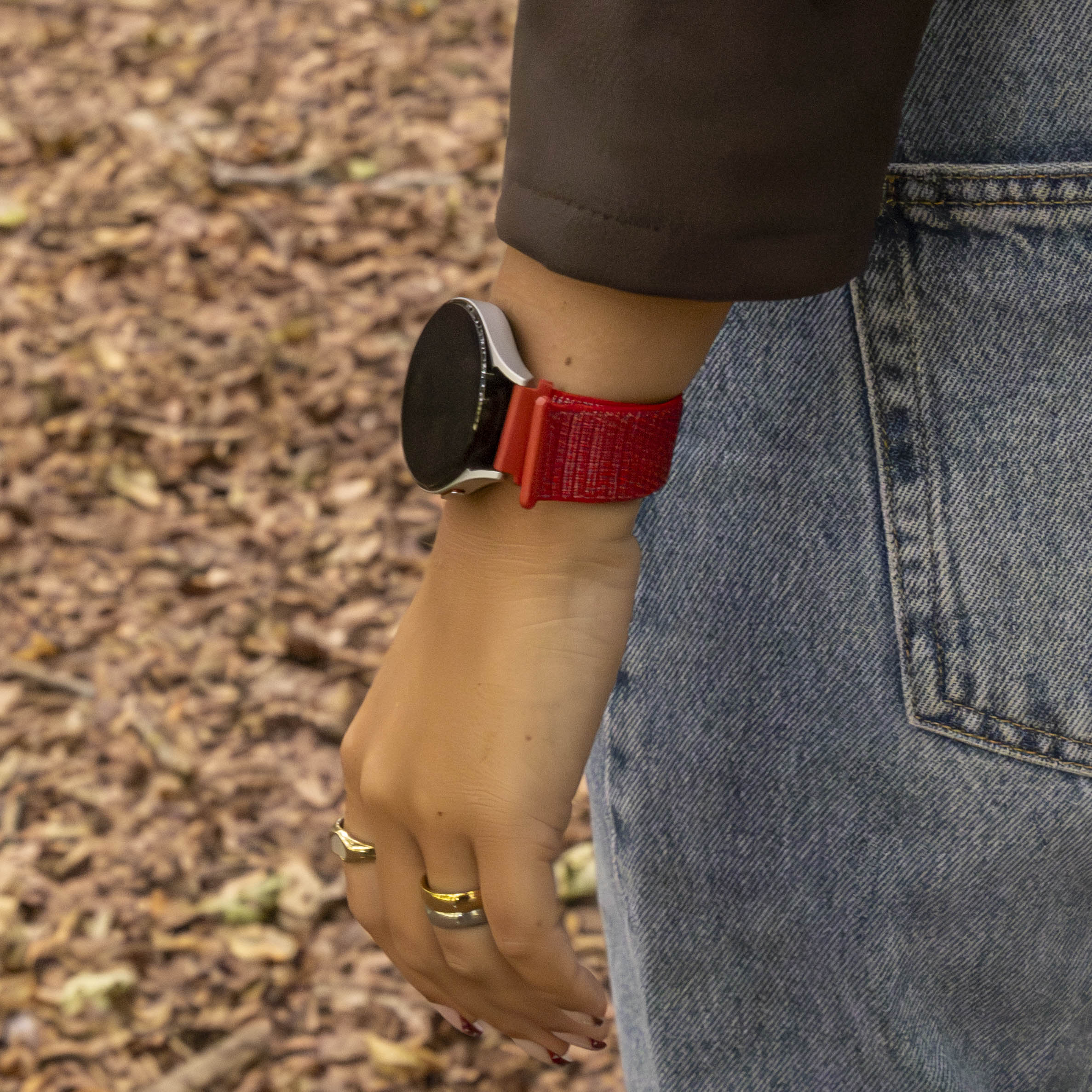 Hand mit Smartwatch und rotem Armband. Person trägt Ringe und Jeans. Verschwommener Hintergrund mit Blättern.