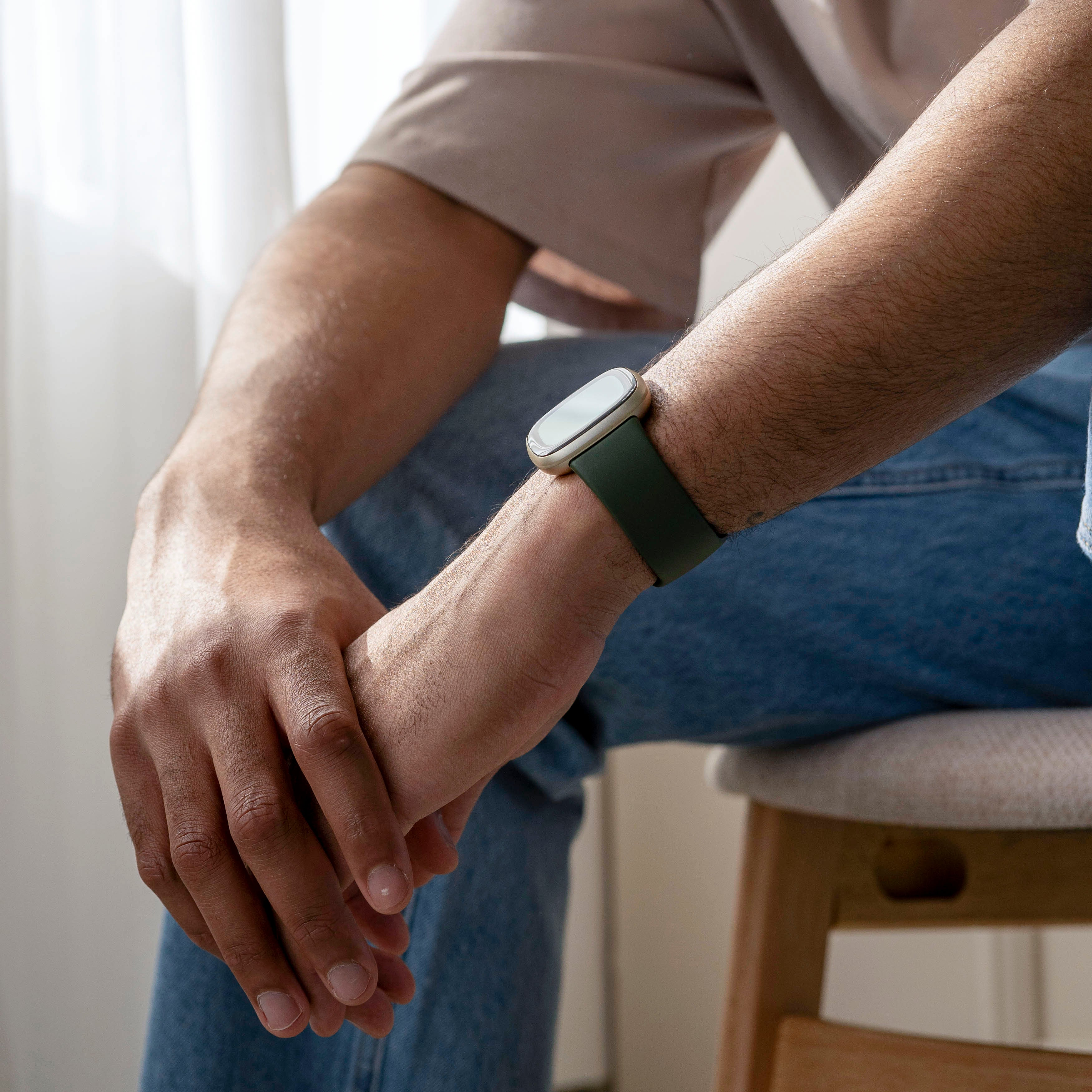 Eine Person sitzt mit verschränkten Armen. Eine Uhr mit grünem Armband am Handgelenk. Beiges Hemd, Jeans.