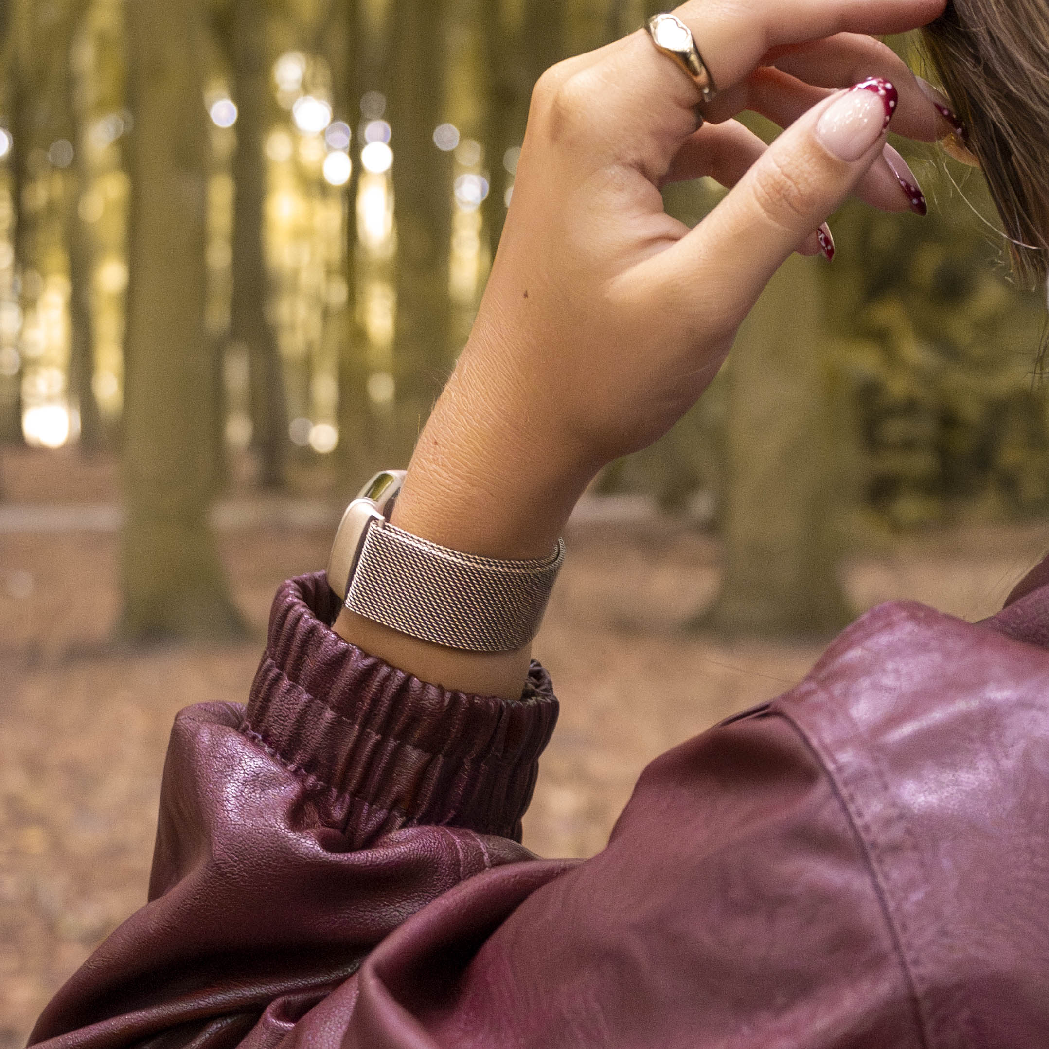 Eine Hand mit Ring und gepunkteten Nägeln berührt die Haare. Eine Uhr befindet sich am Handgelenk. Eine Lederjacke vor Parkhintergrund.