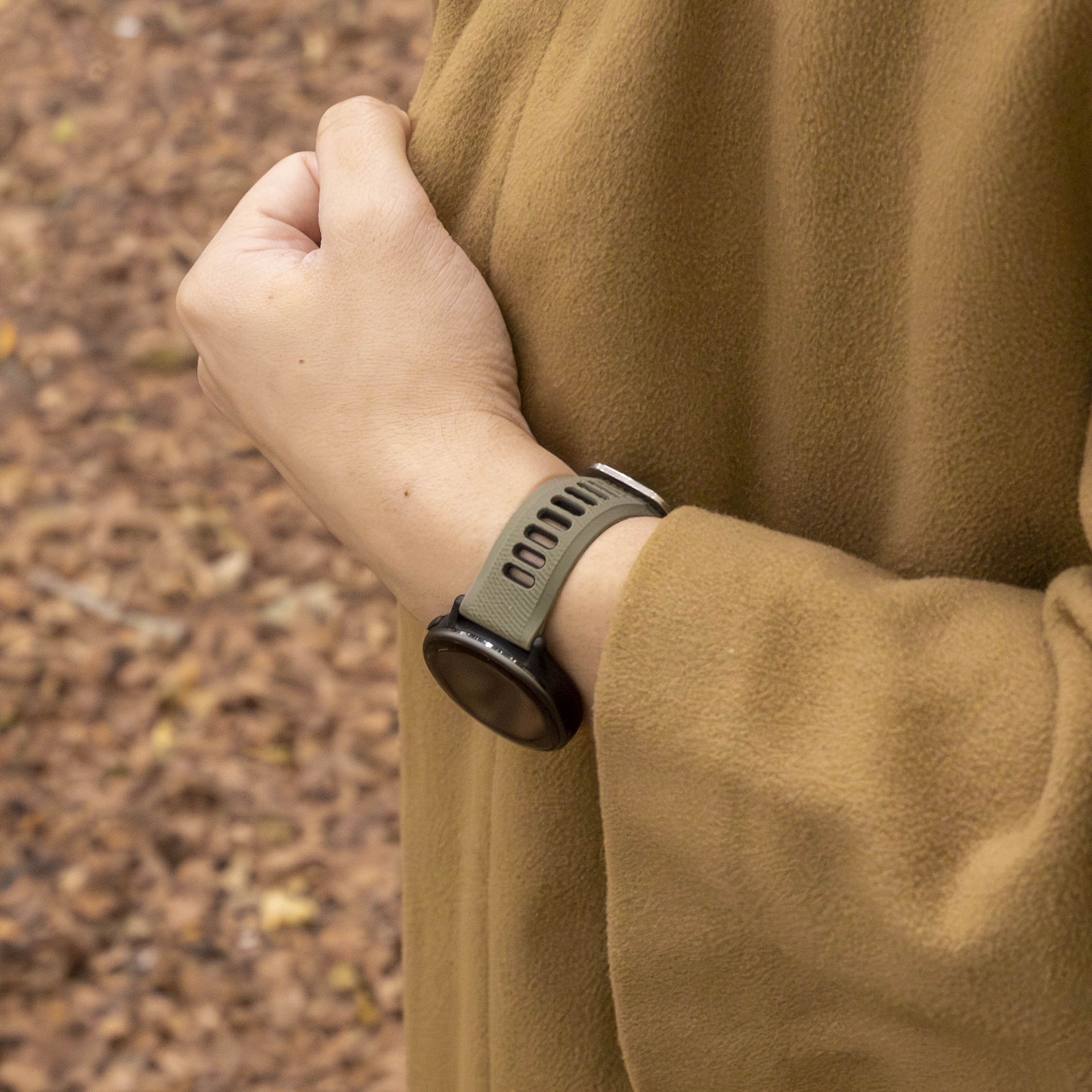 Das Handgelenk einer Person mit einer grünen Uhr. Die Person trägt einen braunen Mantel, der Hintergrund ist unscharf mit braunen Blättern.