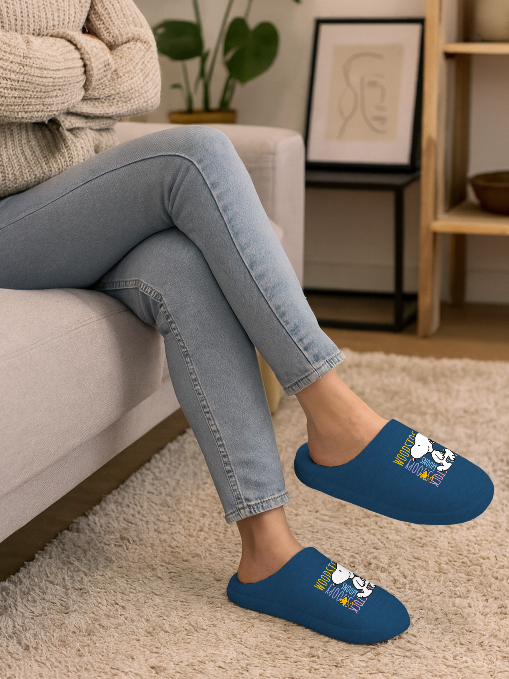 Person in blauen Hausschuhen, sitzt auf einer Couch. Die Hausschuhe haben ein Snoopy-Design. Die Person trägt einen Pullover und Jeans.