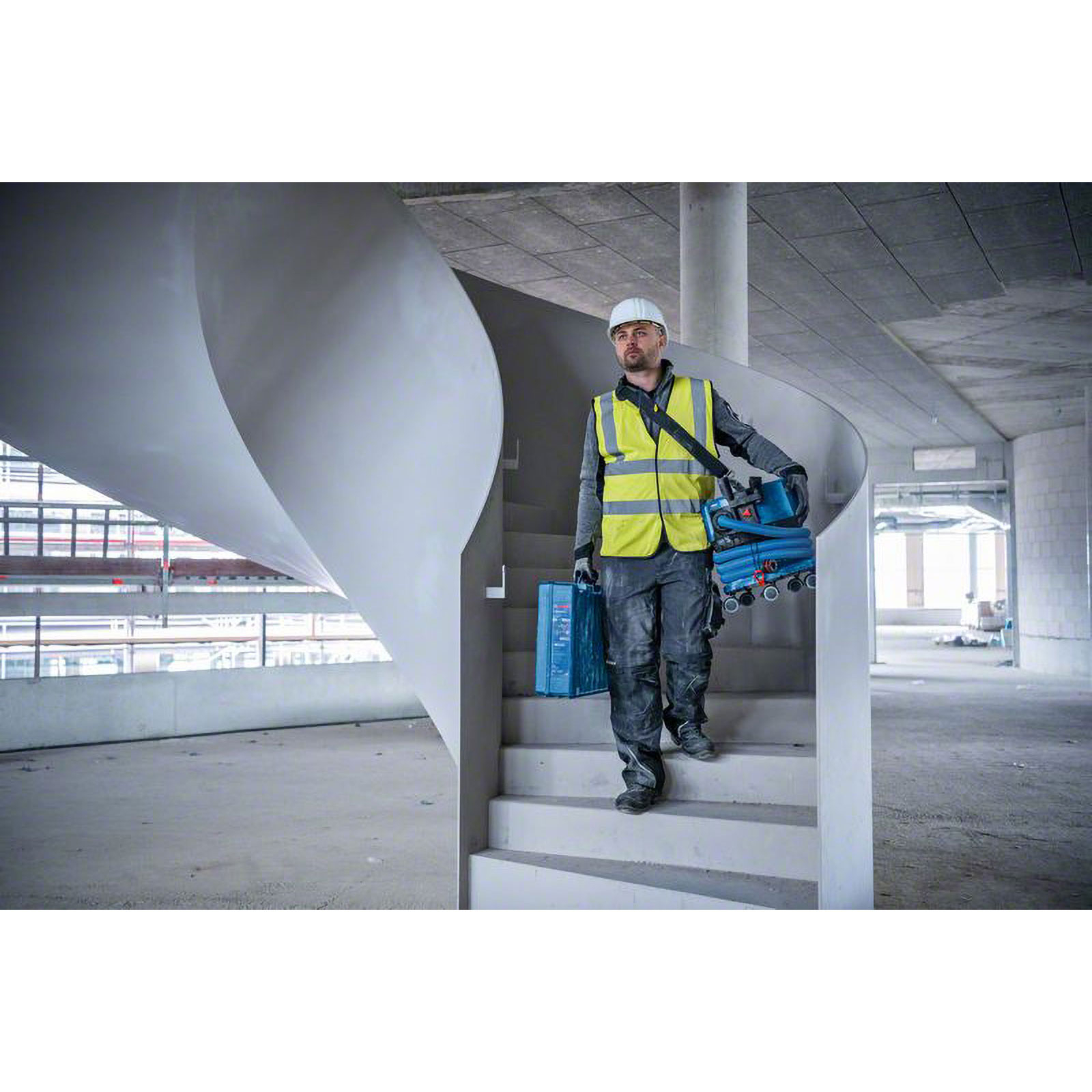 Ein Bauarbeiter mit gelber Weste trägt Werkzeuge in einem Gebäude die Treppe hinunter. Weißer Helm, blauer Werkzeugkoffer und Maschinen.