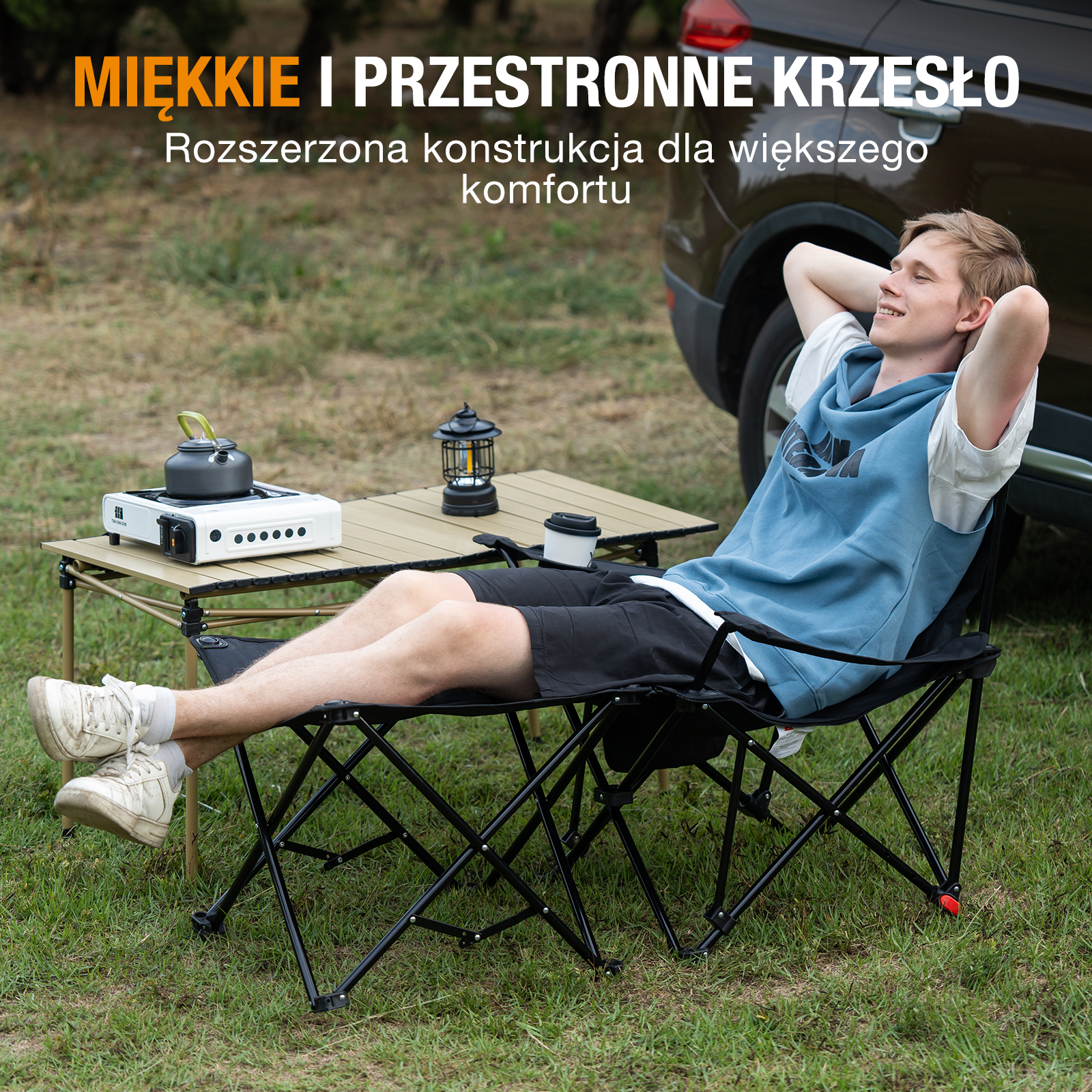 Mężczyzna relaksuje się w czarnym krześle składanym. W pobliżu stół z kuchenką i latarnią. Samochód w tle.