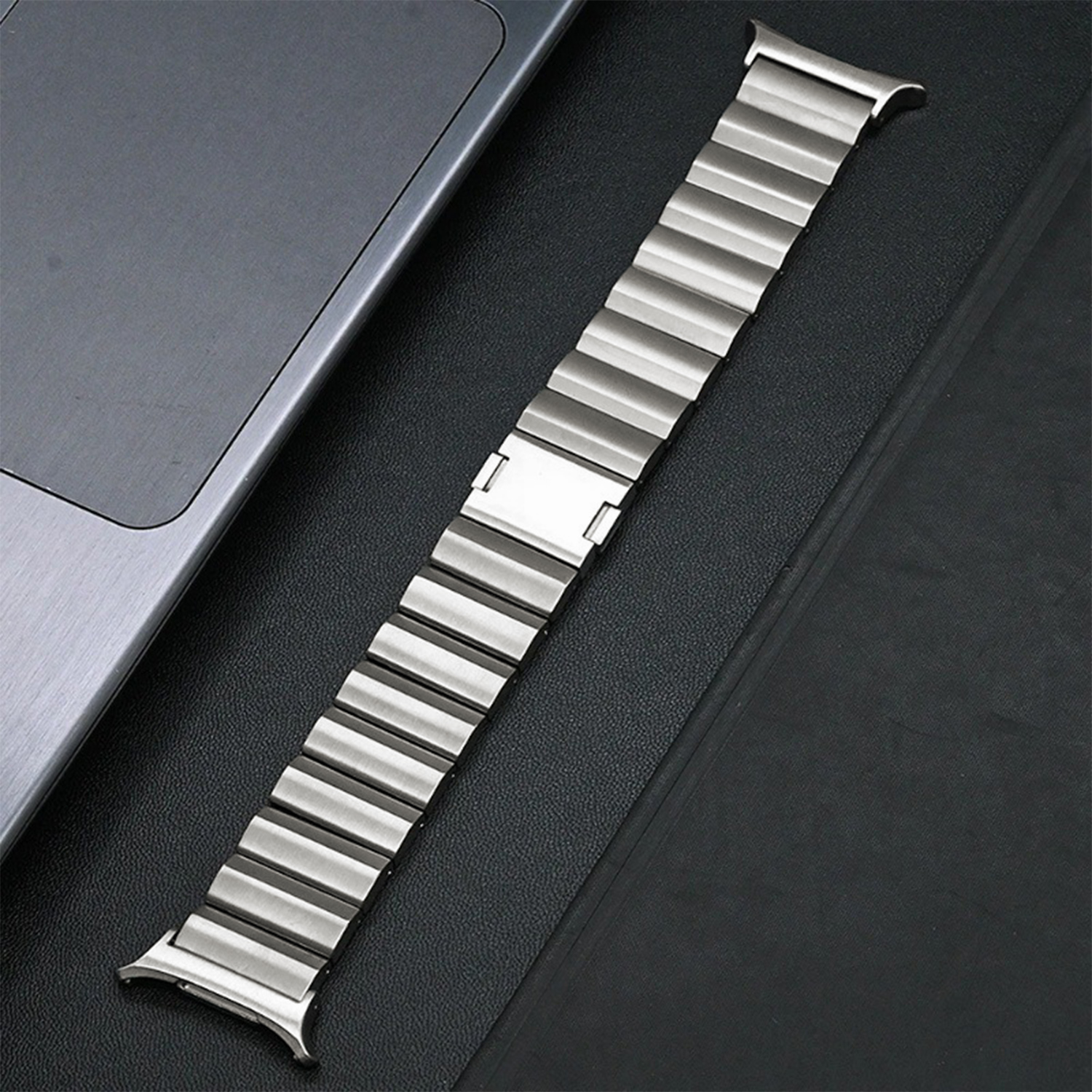 Ein silbernes Uhrenarmband liegt auf einer schwarzen Oberfläche neben einem grauen Laptop. Das Band hat rechteckige Glieder.