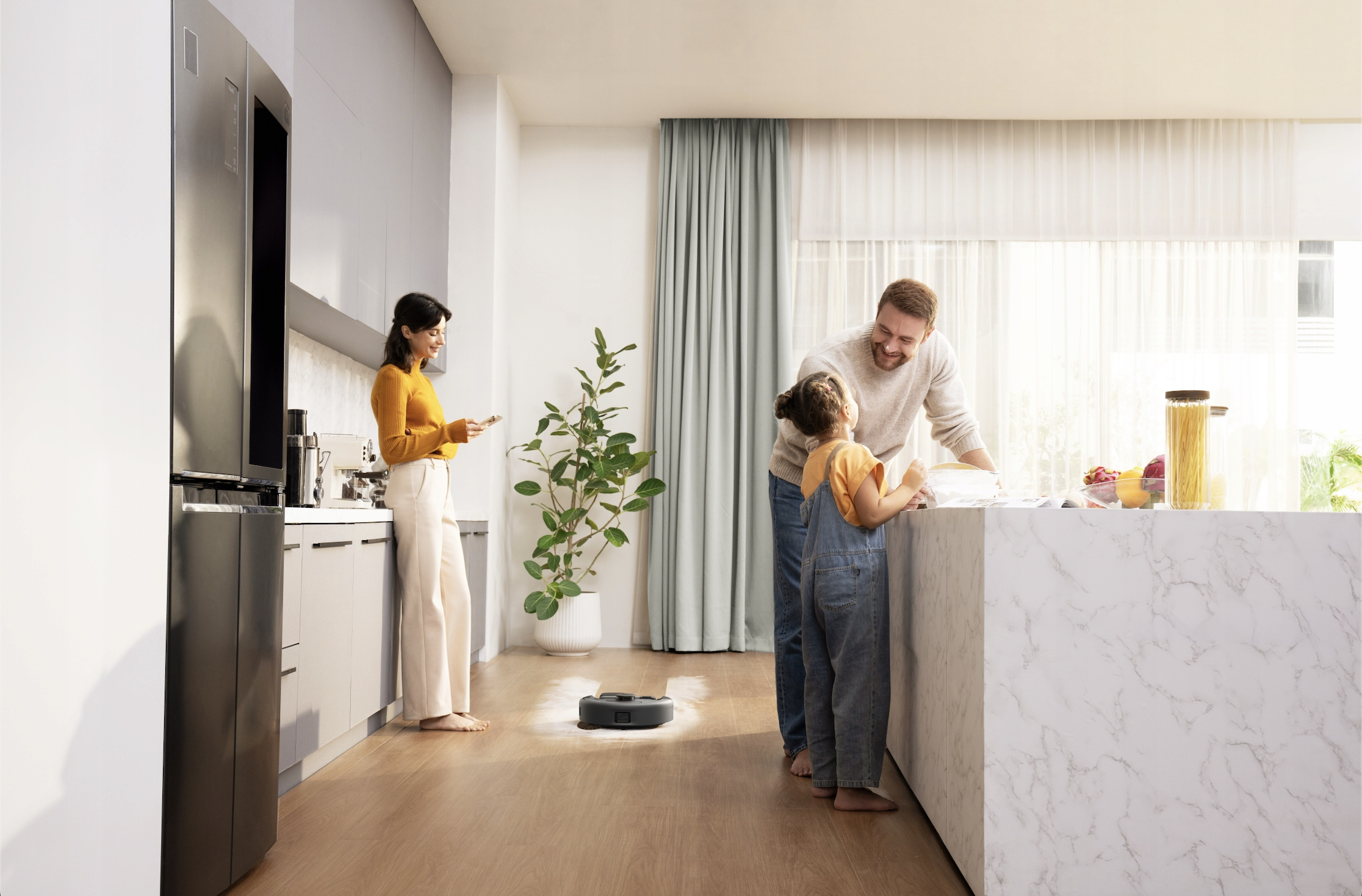 Rodzina w nowoczesnej kuchni. Kobieta po lewej, ojciec i córka przy blacie. Robot sprząta podłogę.