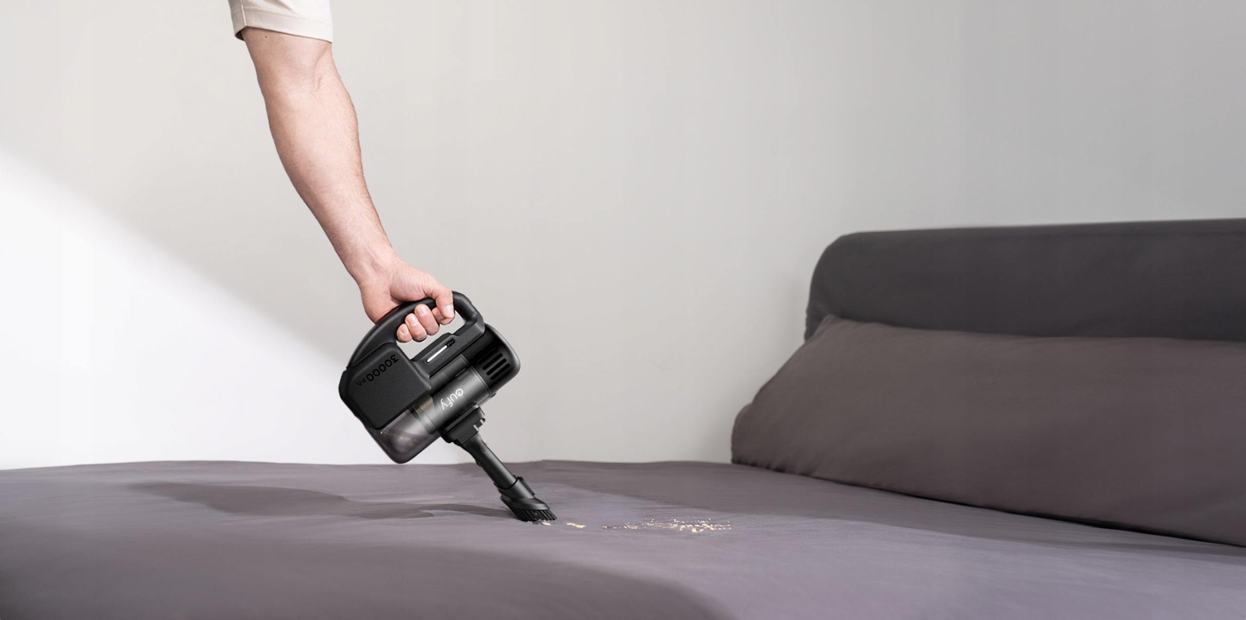 Osoba odkurza łóżko odkurzaczem ręcznym. Łóżko jest szare, a odkurzacz czarny. Na łóżku są jakieś zanieczyszczenia.