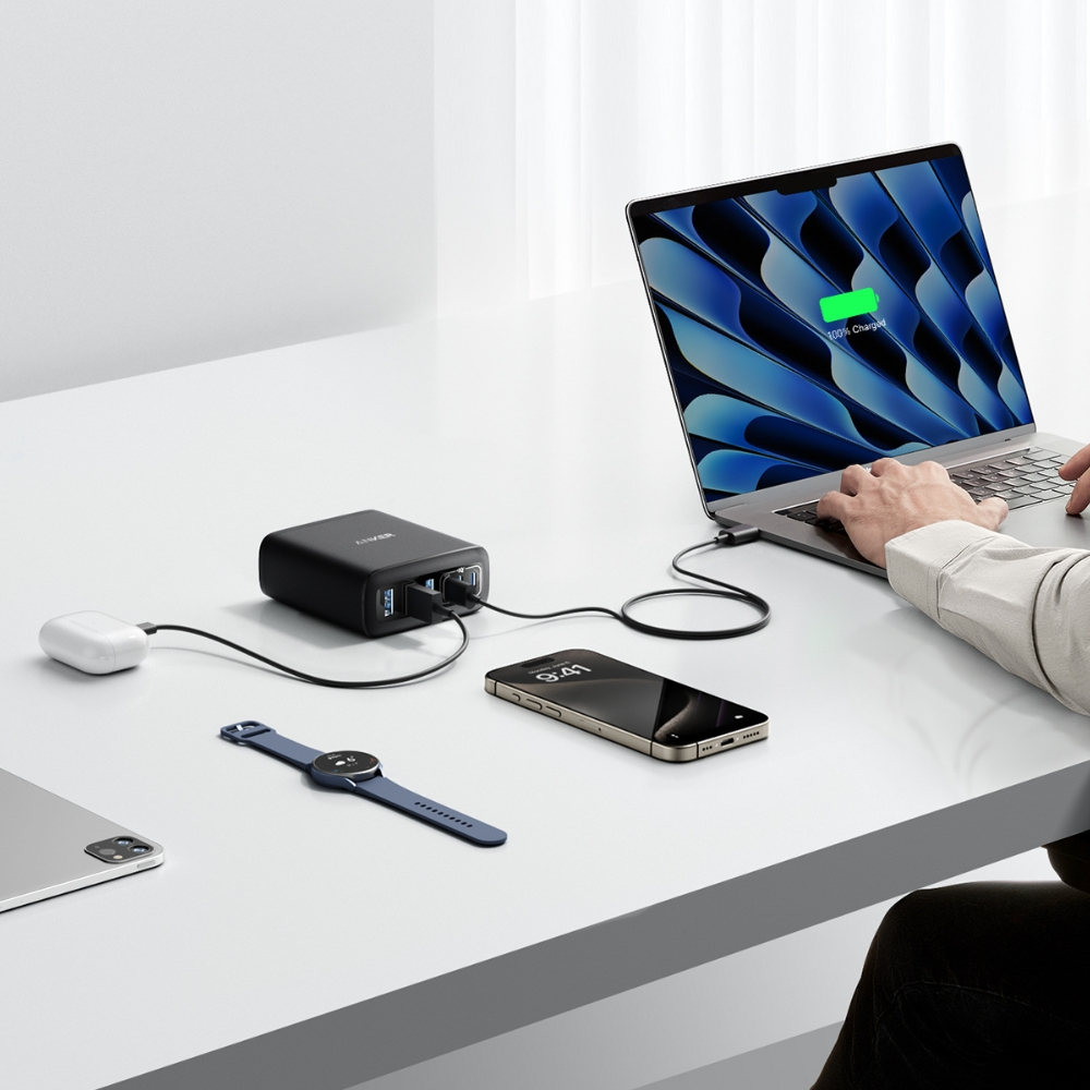Un laptop, un telefono, un orologio e altri dispositivi in carica su una superficie bianca. È presente anche un adattatore di alimentazione. Il laptop è aperto.