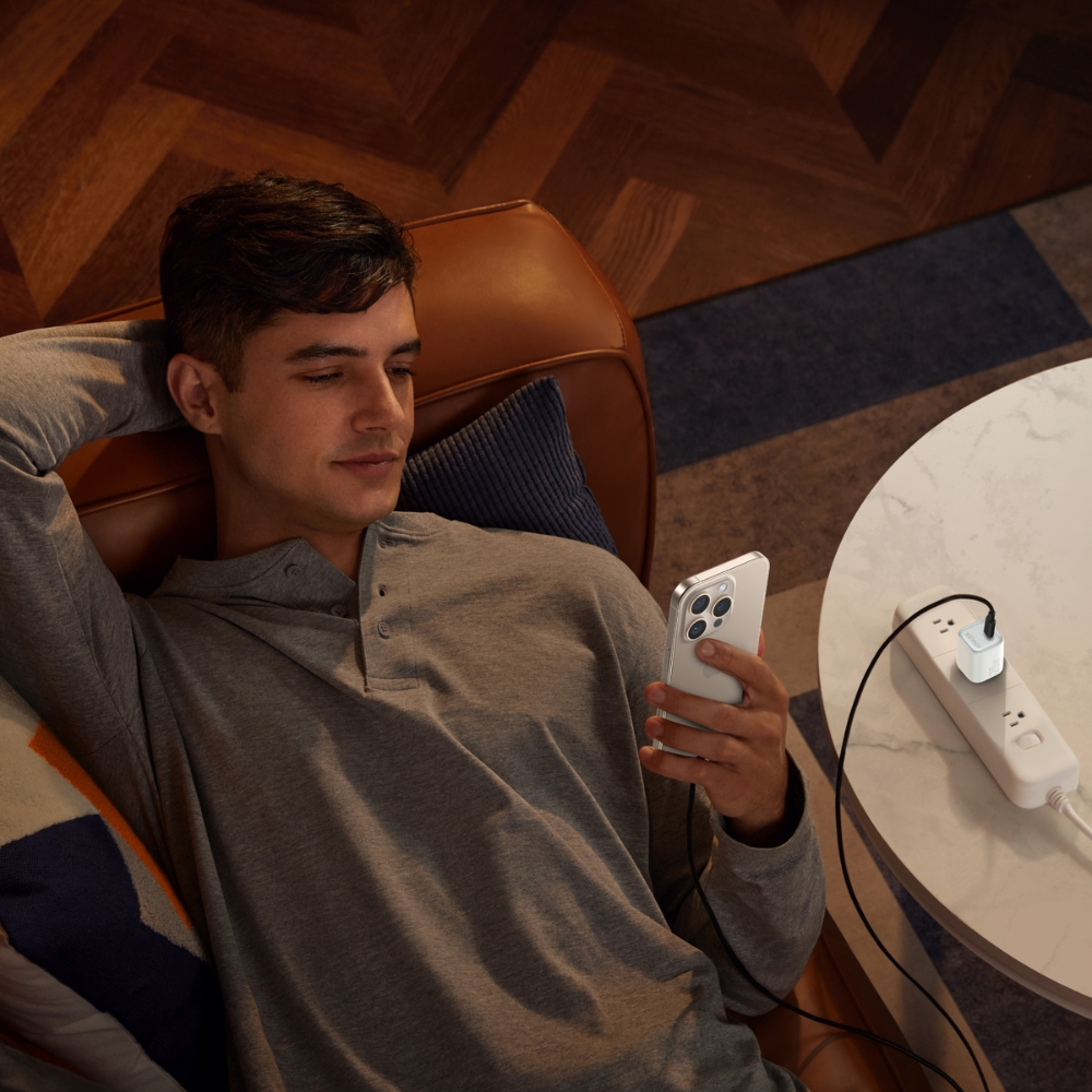 Uomo reclinato, guarda il telefono. Ciabatta sul tavolo. Sedia in pelle marrone, camicia grigia. Pavimento in legno e tappeto fantasia.