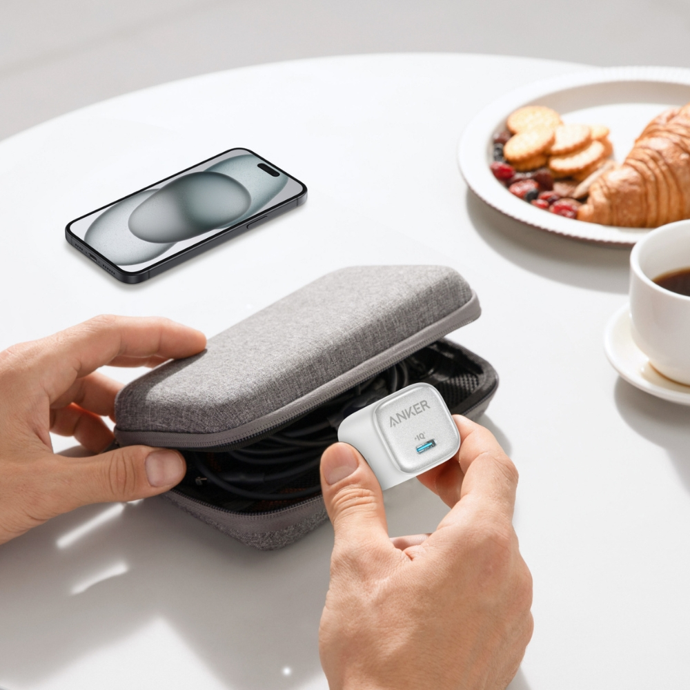 Una persona tiene in mano un caricabatterie Anker bianco. Un telefono, una custodia, articoli per la colazione e una tazza di caffè su un tavolo bianco.