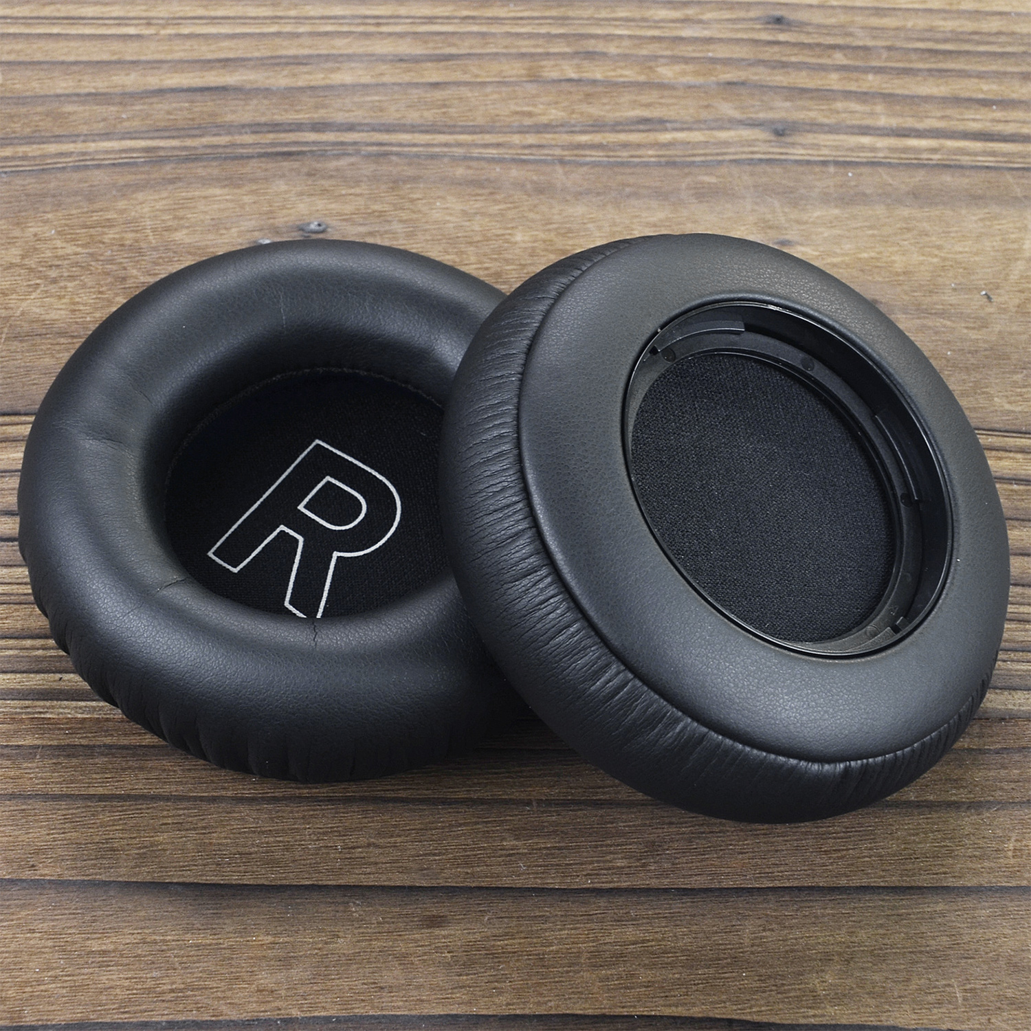 Dwie czarne nausznice do słuchawek na drewnianej powierzchni. Jedna poduszka ma widoczne białe "R". Tło to drewno.