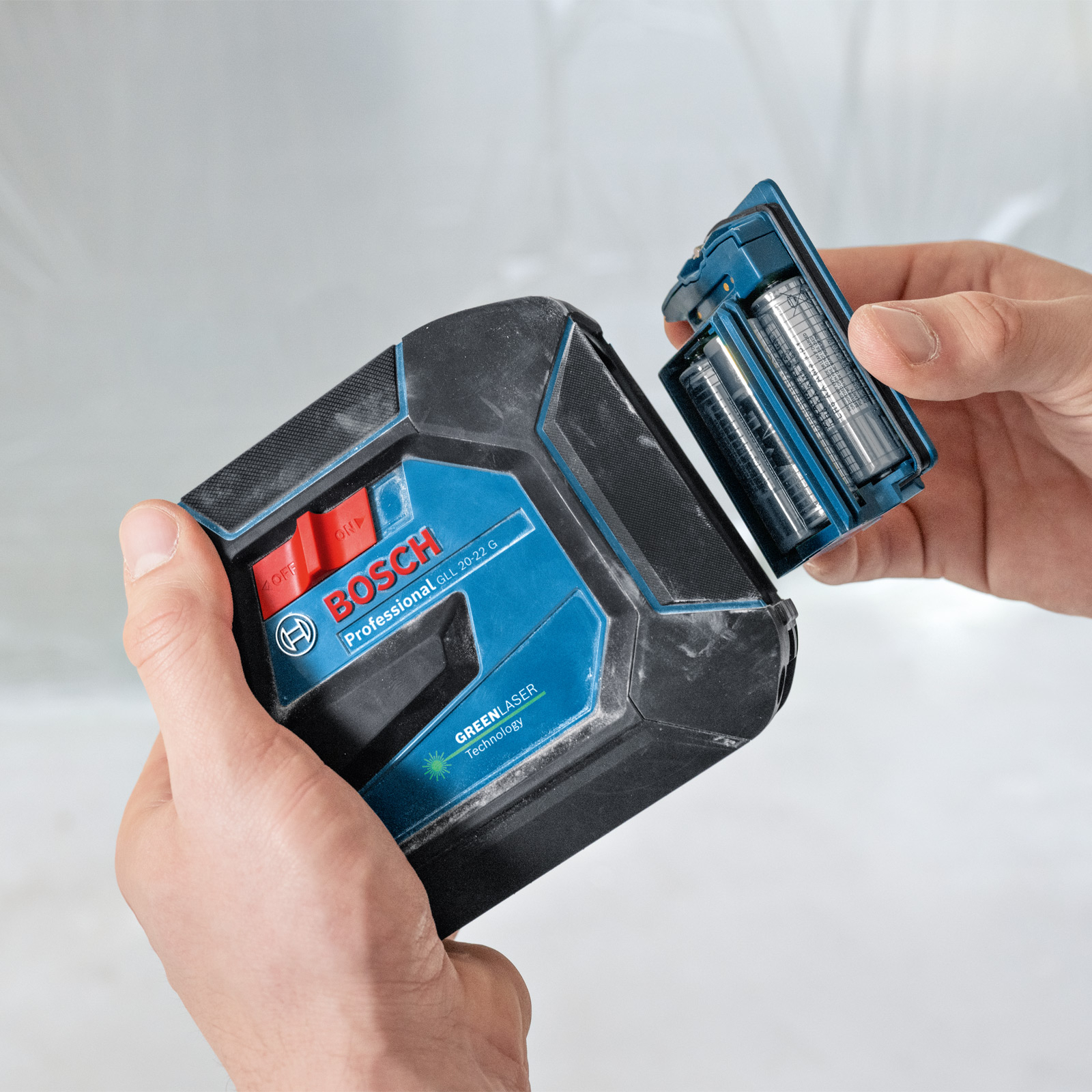 Hände halten ein Bosch-Laser-Niveau und zeigen das Einsetzen der Batterien. Das Gerät ist schwarz und blau.