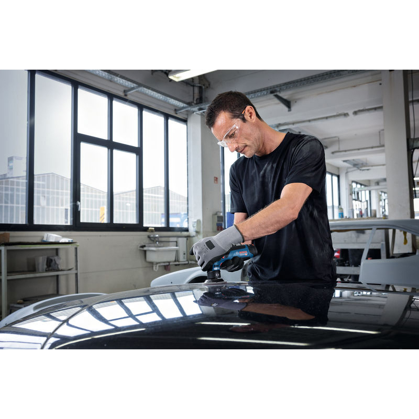 Mann in der Werkstatt, der ein Elektrowerkzeug auf einem Autodach benutzt. Er trägt eine Schutzbrille und Handschuhe.