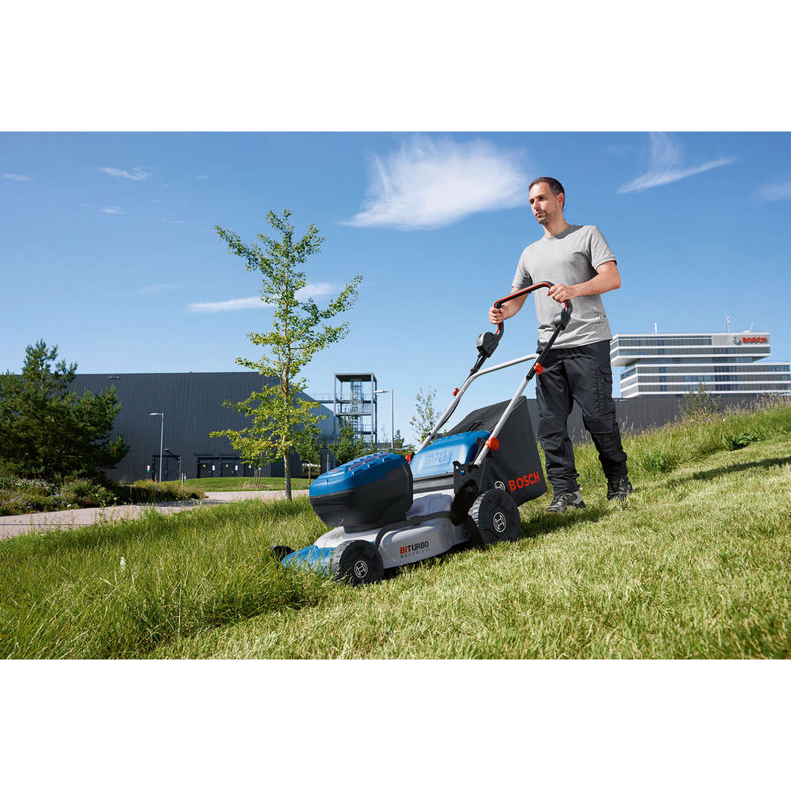 Un hombre corta el césped con un cortacésped eléctrico Bosch en un día soleado, con un edificio al fondo.