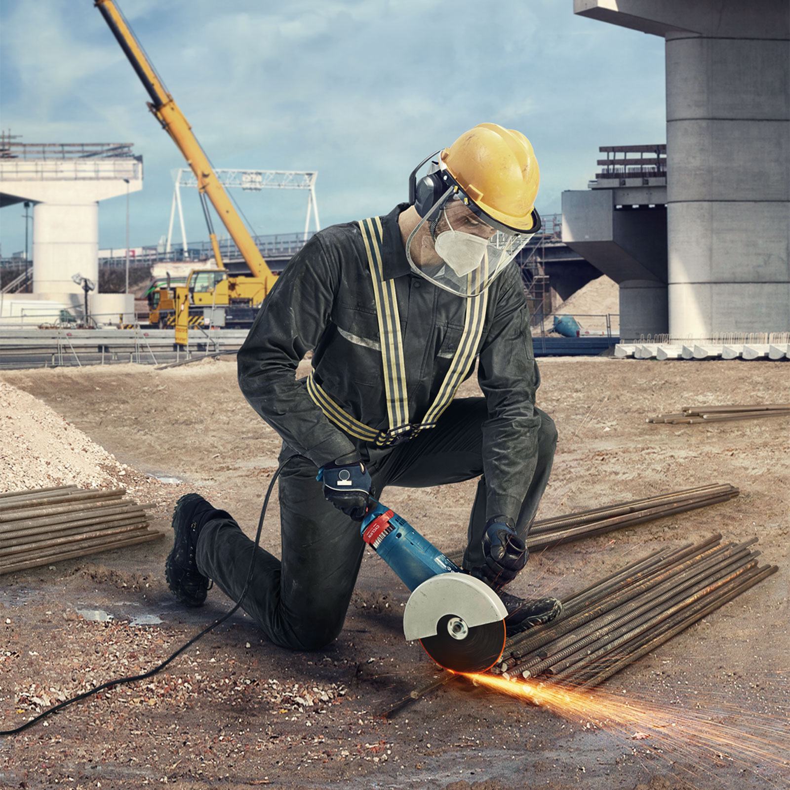 Robotnik klęczy, szlifując pręty zbrojeniowe elektronarzędziem. Iskry lecą. Plac budowy z dźwigiem i mostem w tle.