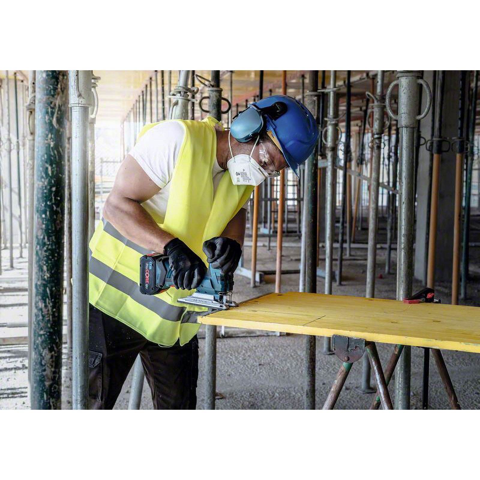 Un operaio edile con dispositivi di sicurezza taglia legno con un seghetto alternativo. La scena è un cantiere con impalcature metalliche.