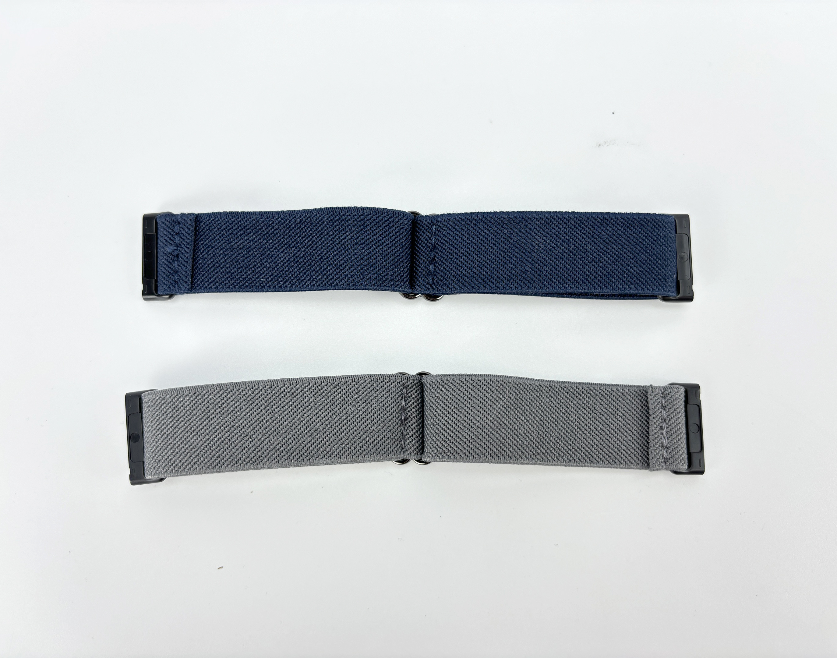 Zwei Uhrenarmbänder: eines marineblau, eines grau. Beide haben schwarze Verschlüsse und liegen auf weißem Untergrund.