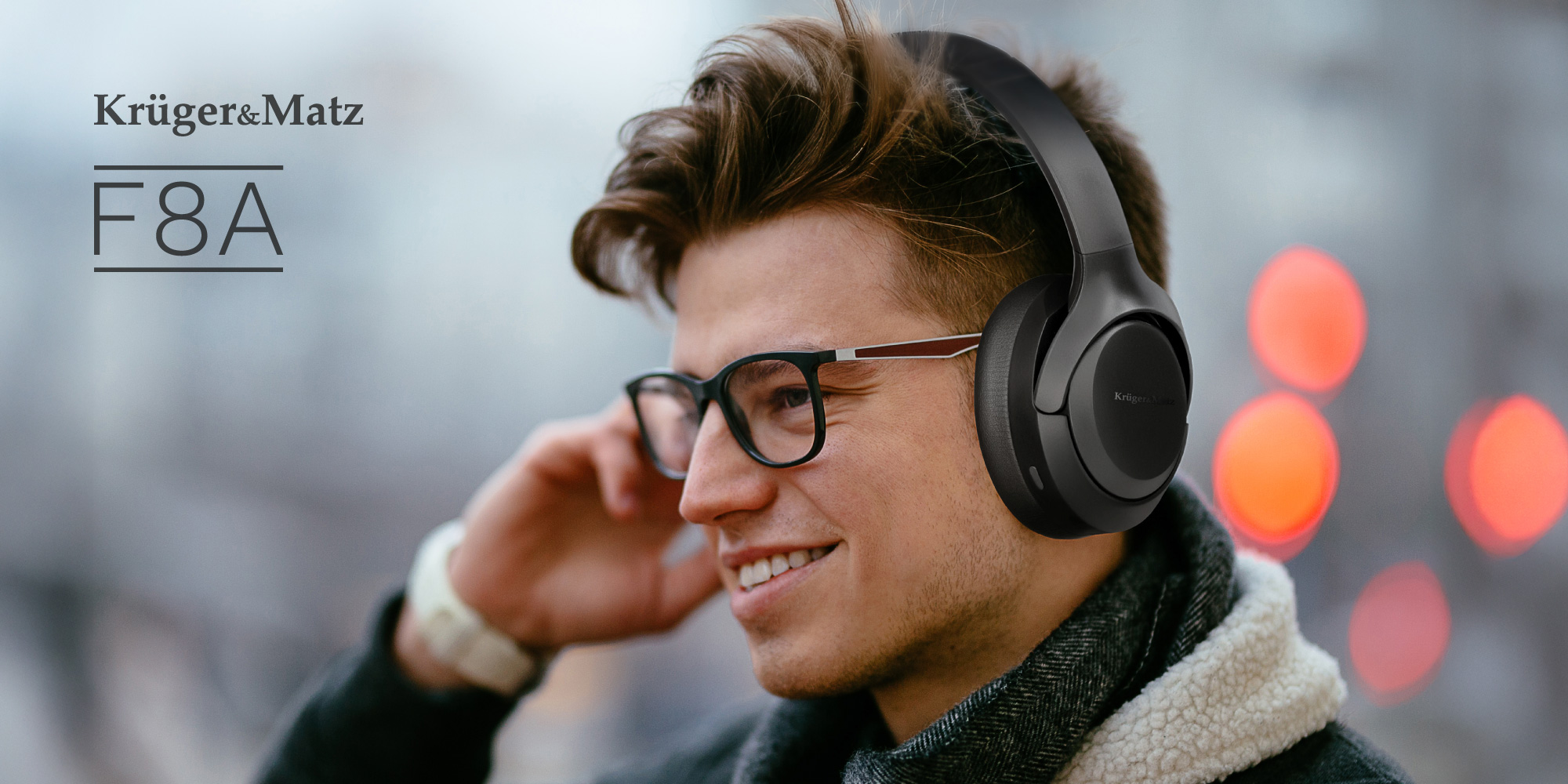 Mężczyzna w okularach i czarnych słuchawkach uśmiecha się na zewnątrz. Obraz przedstawia reklamę produktu z logo 'Kruger&Matz F8A'.