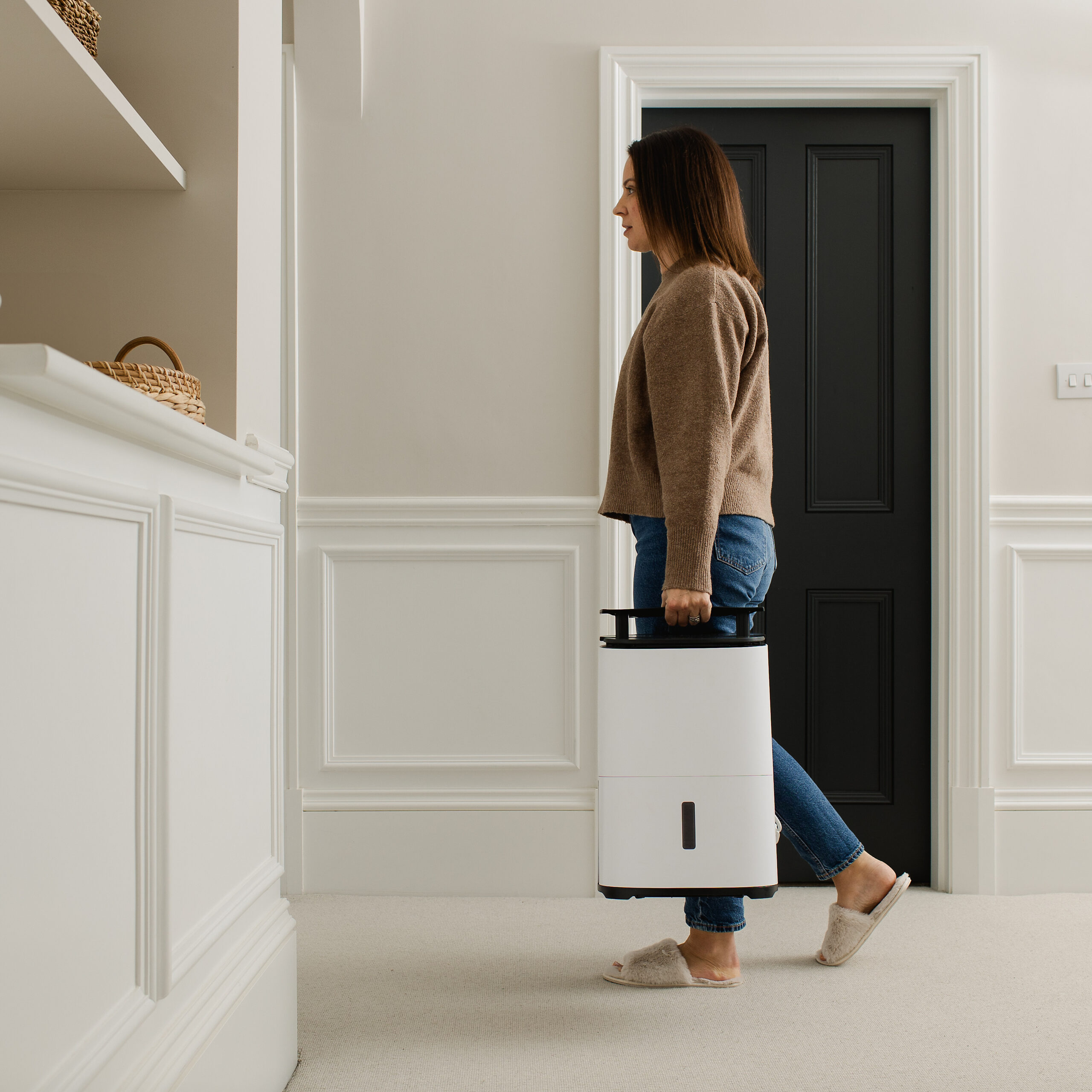 Kobieta w korytarzu niosąca biały osuszacz powietrza. Ma na sobie brązowy sweter, dżinsy i kapcie. Widoczne czarne drzwi.