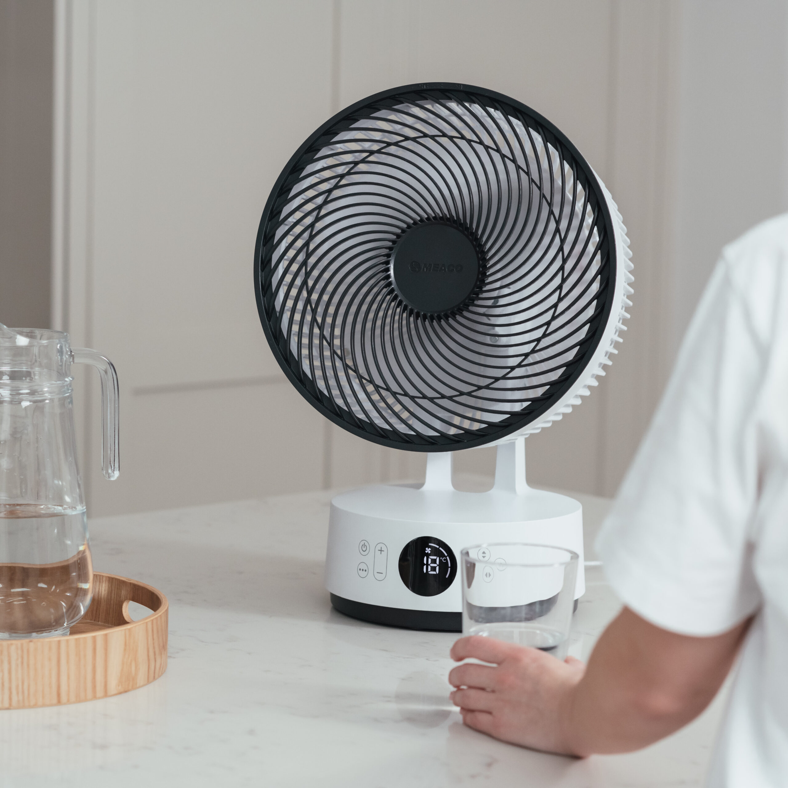 Biały wentylator z czarnymi łopatkami stoi na blacie. Osoba trzyma szklankę wody. Dzbanek z wodą jest po lewej stronie.