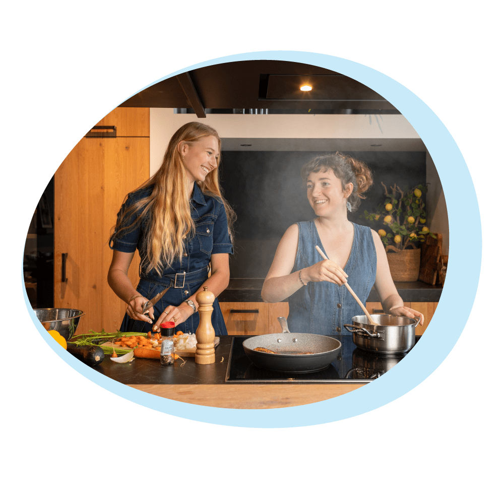 Zwei Frauen kochen in einer Küche. Eine schneidet Gemüse, die andere rührt in einem Topf. Beide tragen Jeanskleider und lächeln.