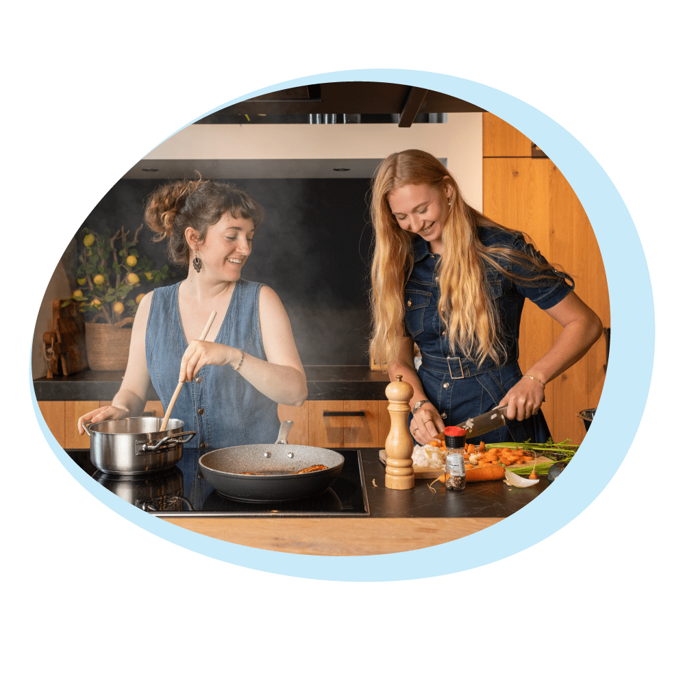 Due donne in cucina, che cucinano. Una mescola in una pentola, l'altra taglia le verdure. La cucina ha mobili in legno ed elettrodomestici in acciaio inox.