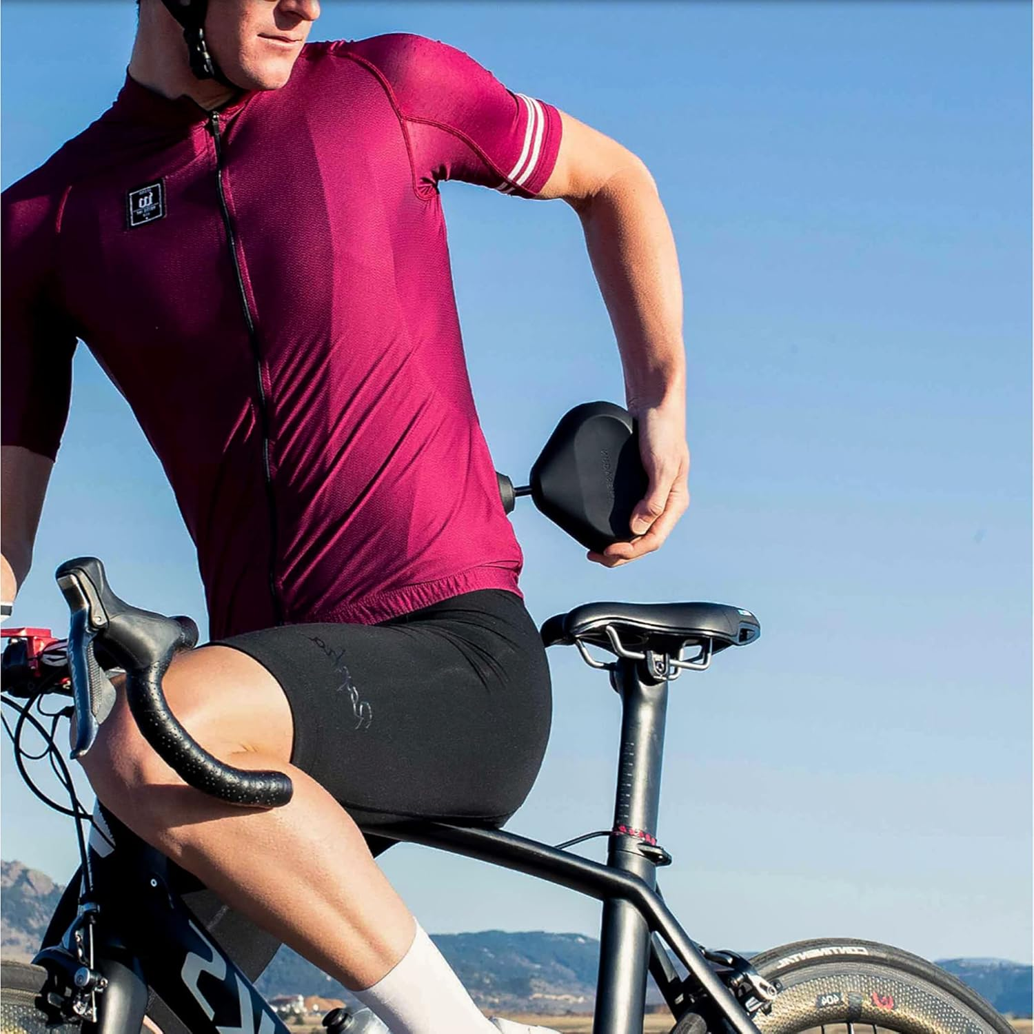 Ciclista con camiseta granate, pantalones cortos negros, en bicicleta, sosteniendo un objeto negro. Cielo azul, montañas al fondo.