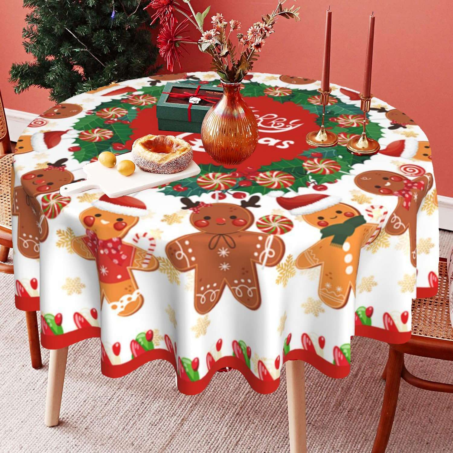 Ein runder Tisch mit einer weihnachtlichen Tischdecke mit Lebkuchenmännern, Süßigkeiten und einem Kranz. Kerzen und eine Vase stehen ebenfalls auf dem Tisch.